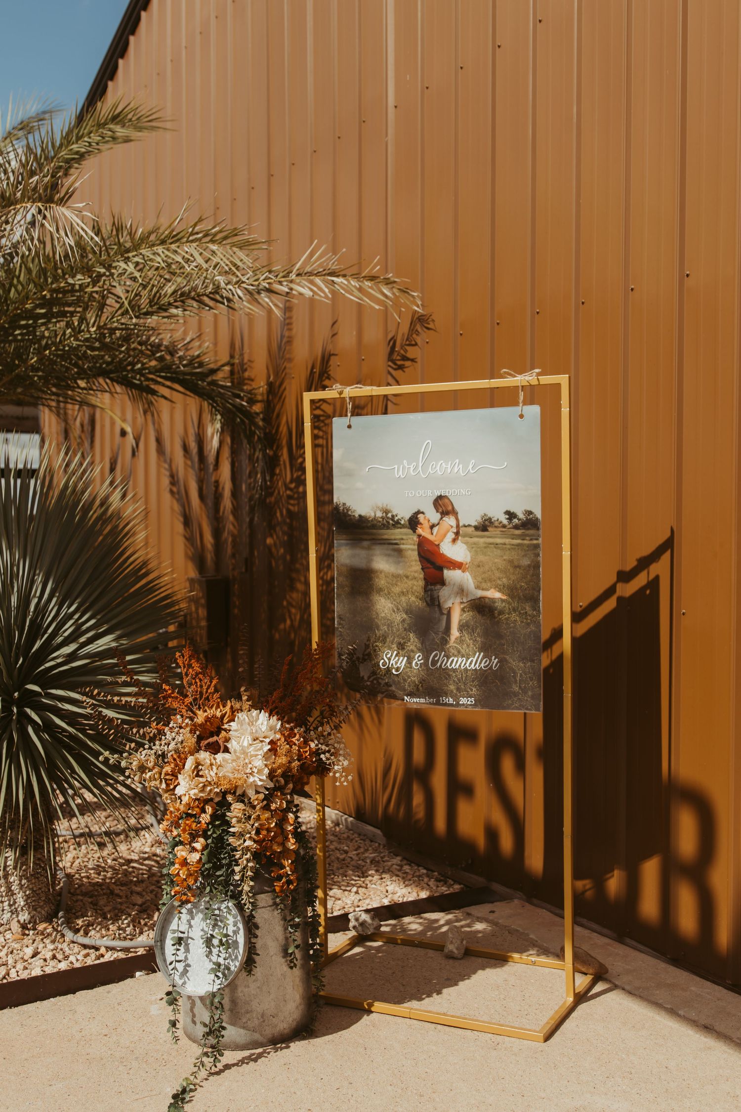 Wedding decor setup featuring palm trees and framed photos against a tan wall with rustic floral arrangements. Wedding photographer DFW