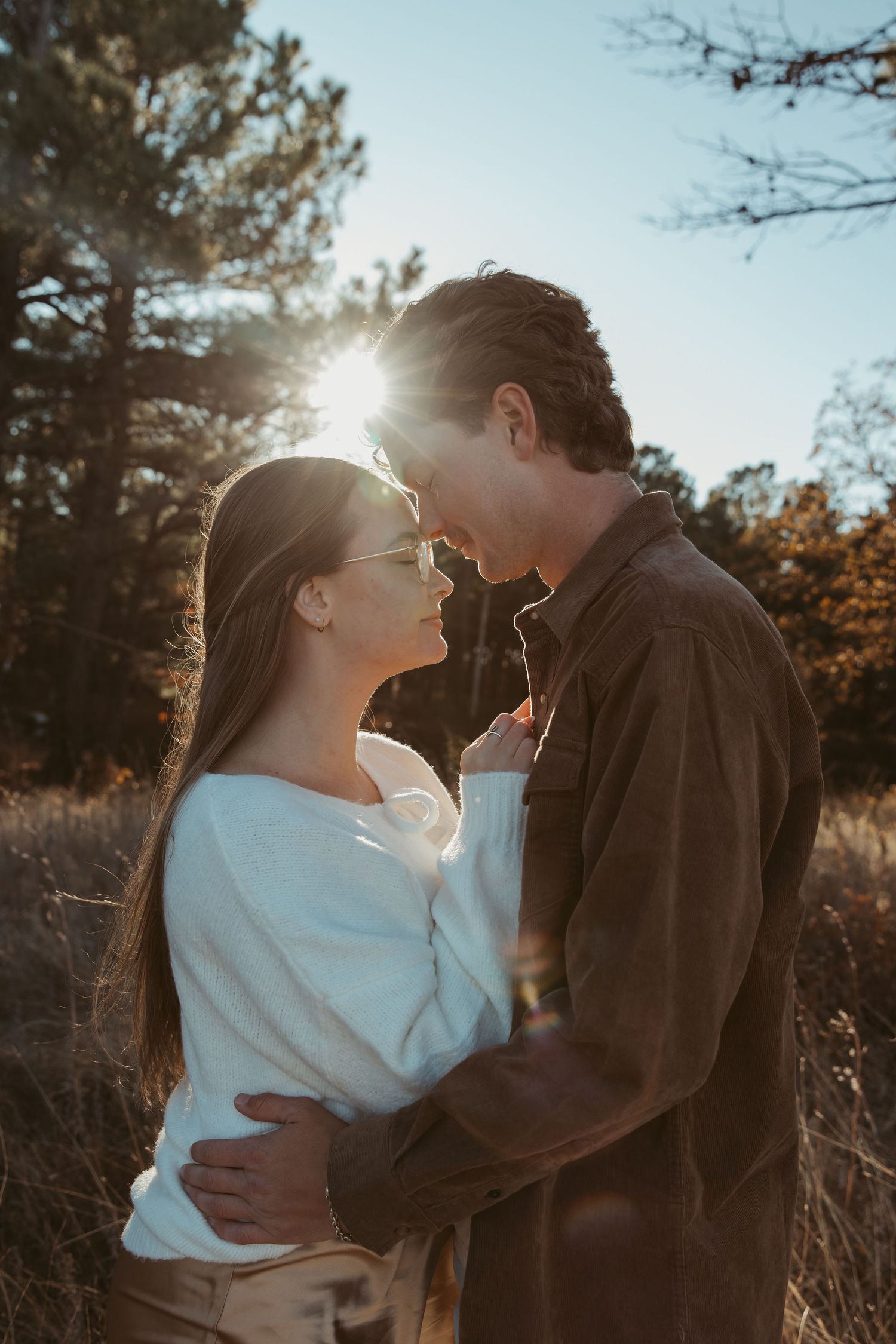 A romantic couple embraces in a sunlit autumn field with golden light streaming through trees in the background. engagement photos DFW
