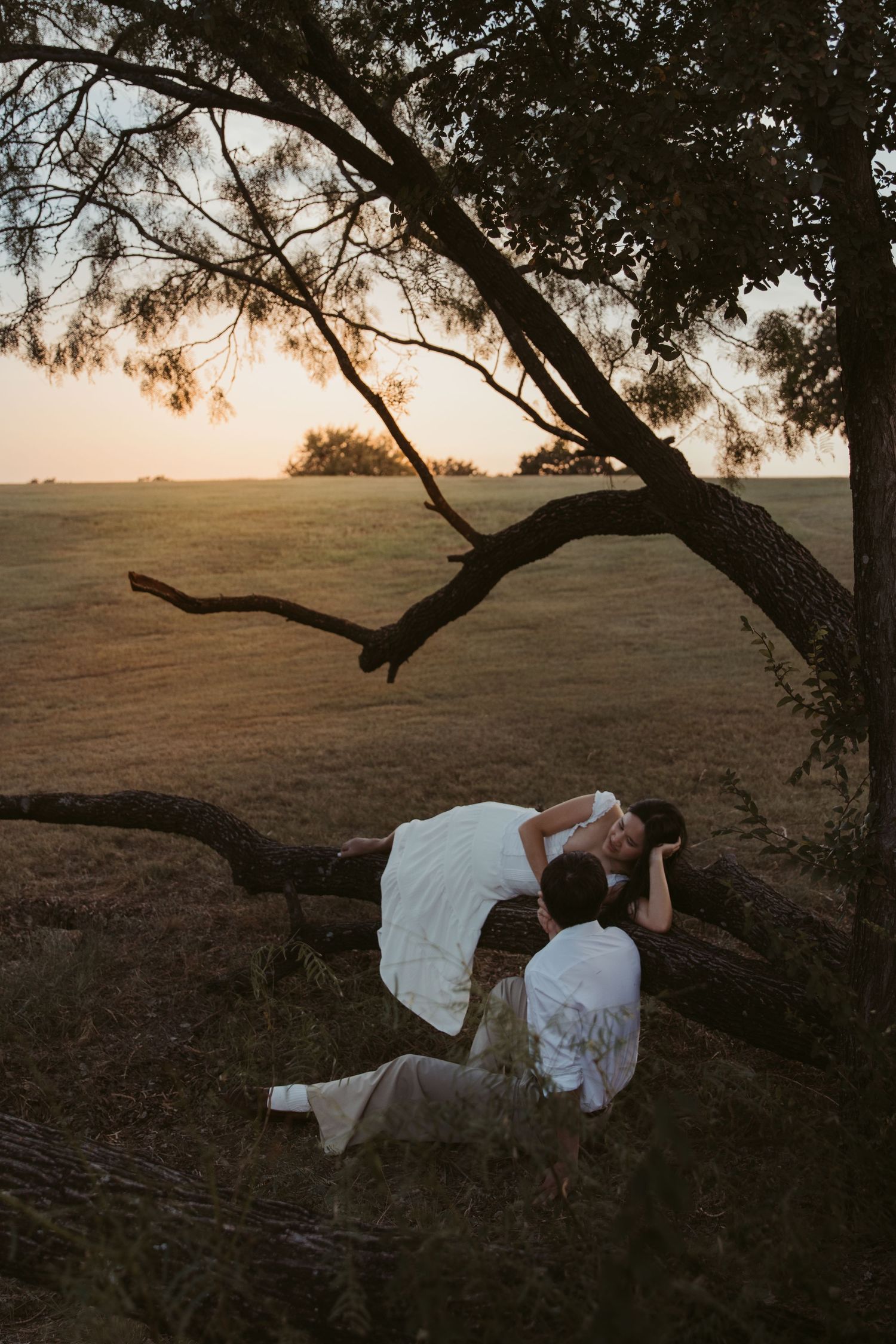 White-dressed figures pose creatively under a twisted tree branch at sunset in a field.