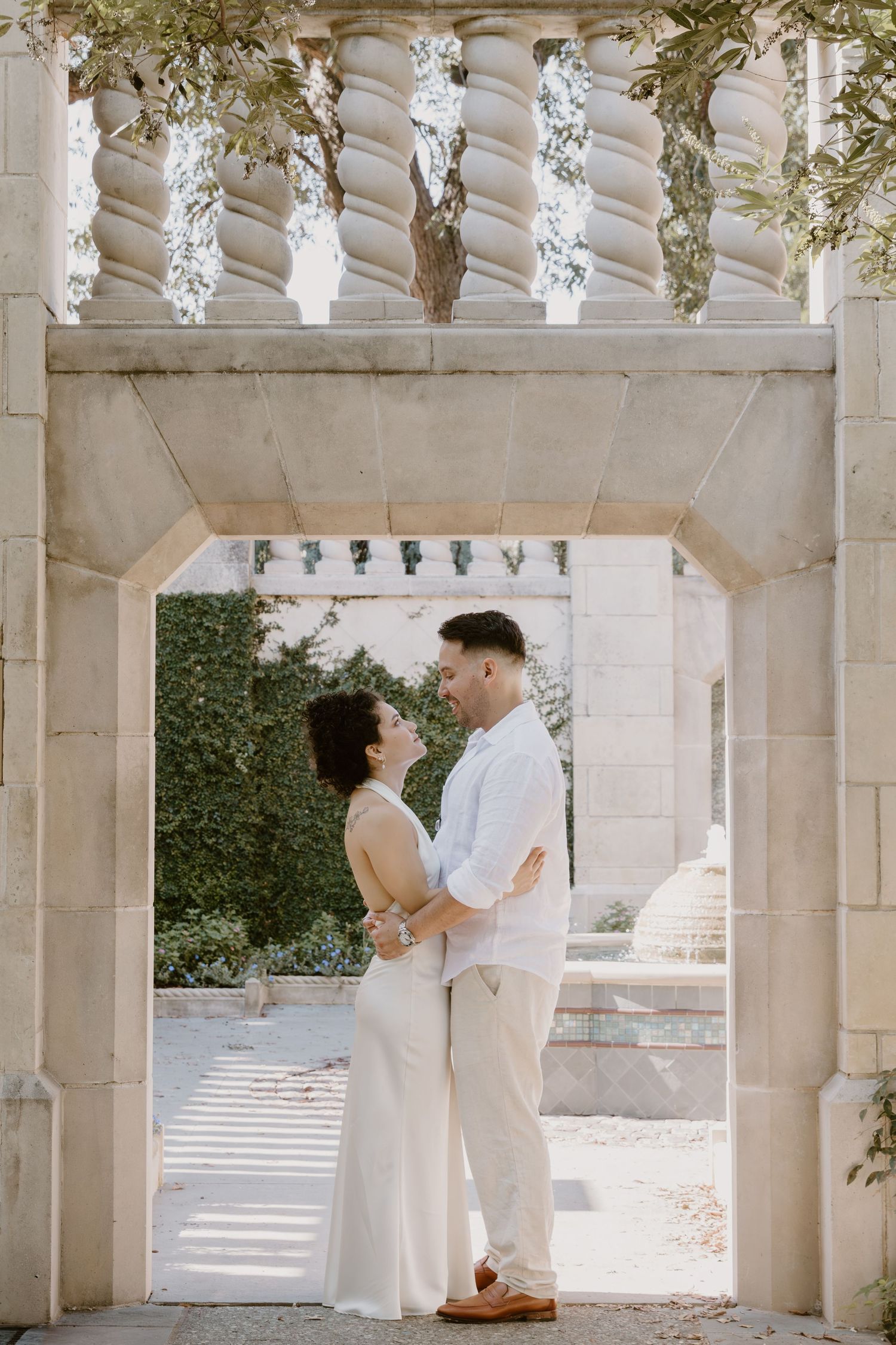 A couple in white clothing embraces under an elegant stone colonnade with hanging decorations.
