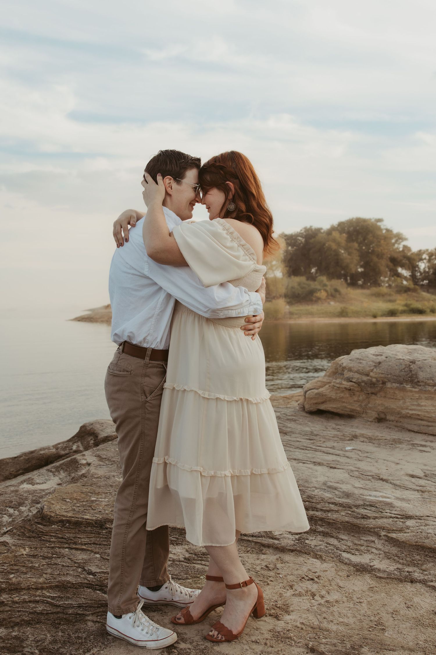 Two people in light colored clothing embrace on rocky shoreline during sunset.