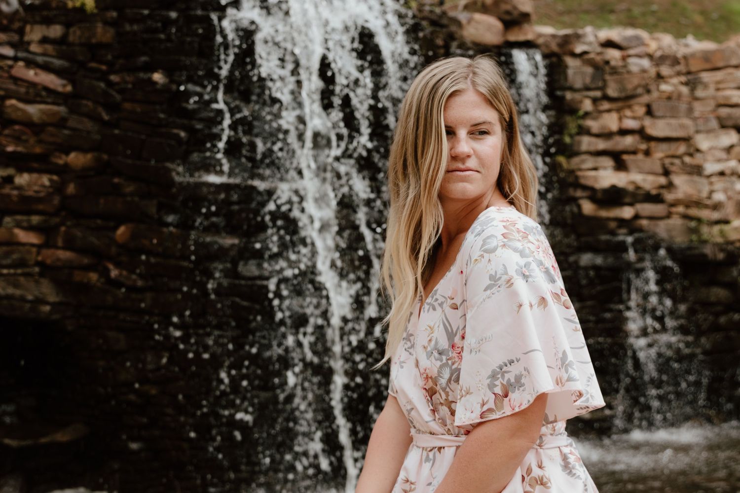 A floral dress catches movement against a stone waterfall backdrop in a romantic outdoor portrait.
