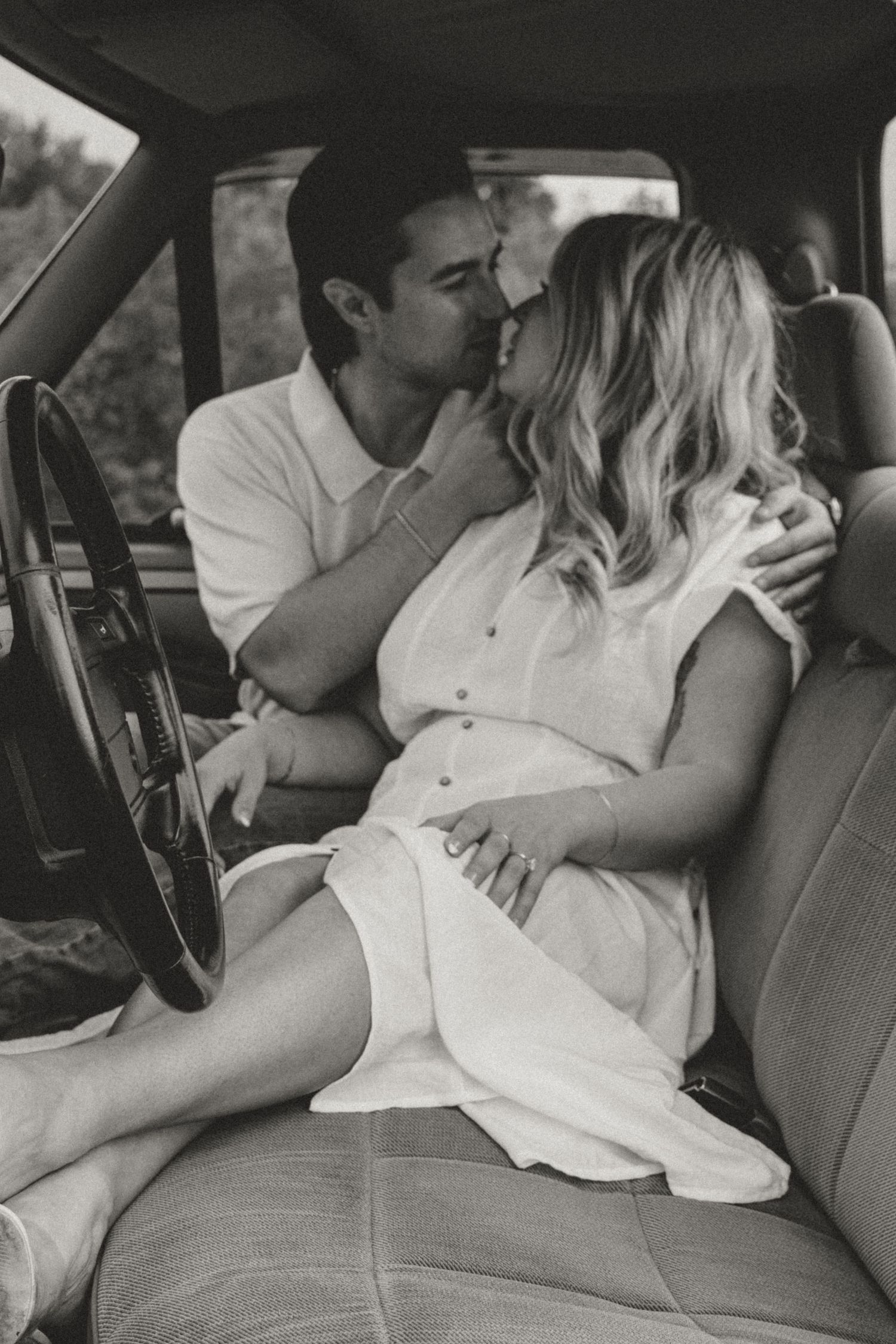 A romantic black and white photograph captures an intimate moment between a couple embracing in the front seat of a vintage car.