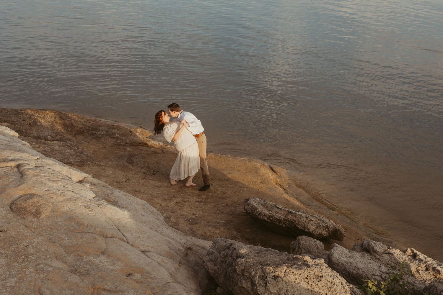 A couple in white attire sharing an intimate moment on rocky cliffs overlooking a serene lake.