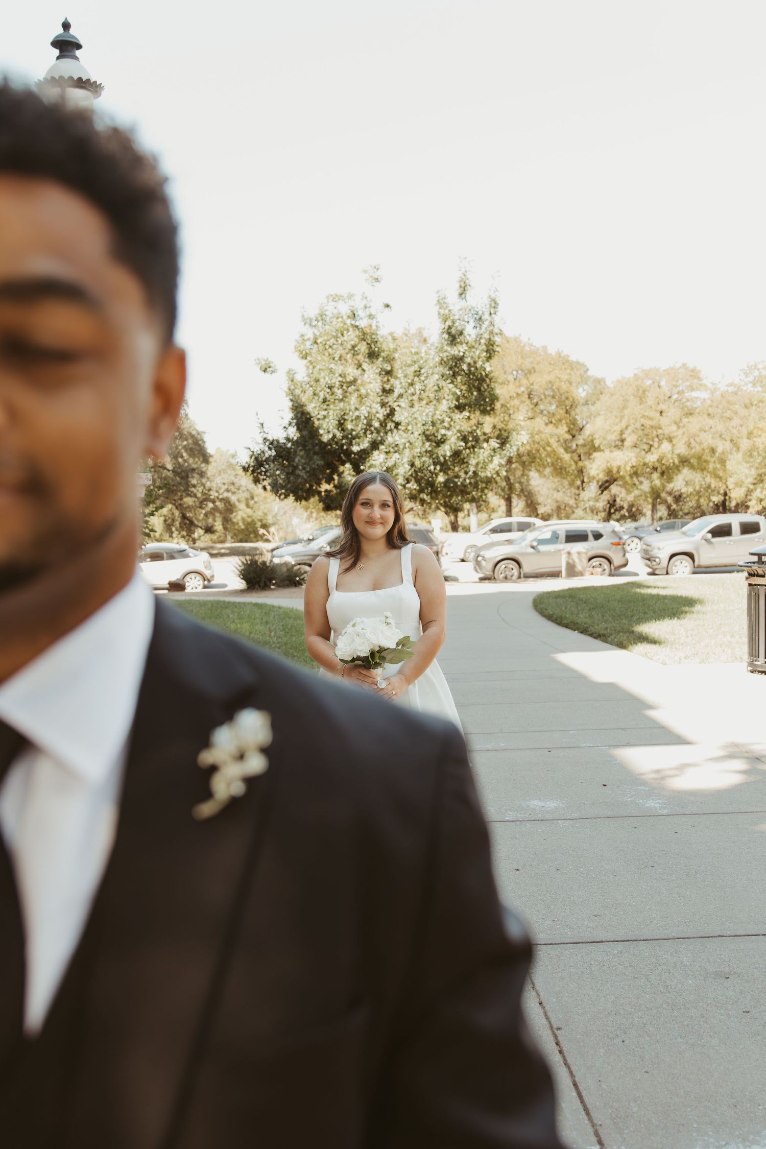 A wedding couple embracing outdoors in a parking lot with trees and cars in the background. Wedding Photographer DFW