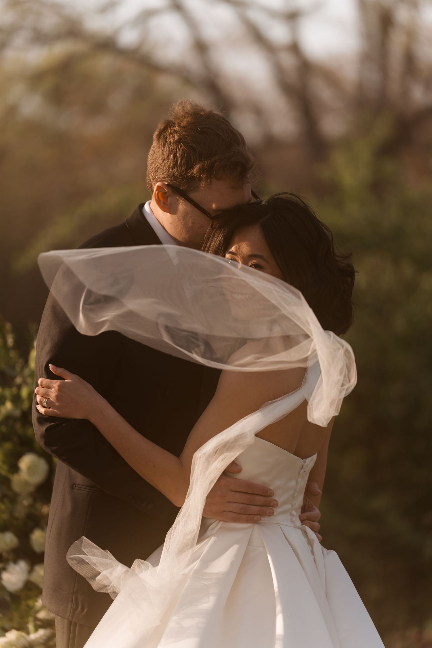 A bride and groom embrace outdoors, her veil flowing in the wind, bathed in warm golden sunlight.