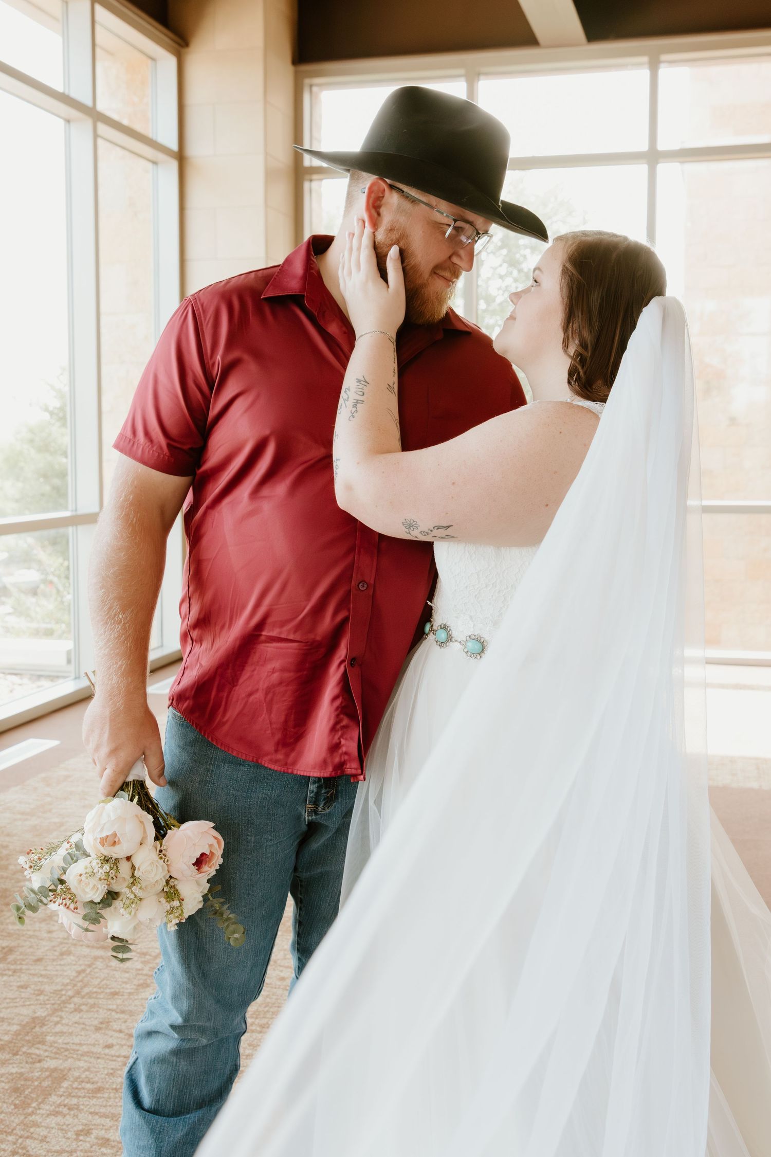 A tender moment between a couple on their wedding day with the bride wearing white and groom in a red shirt and black hat.