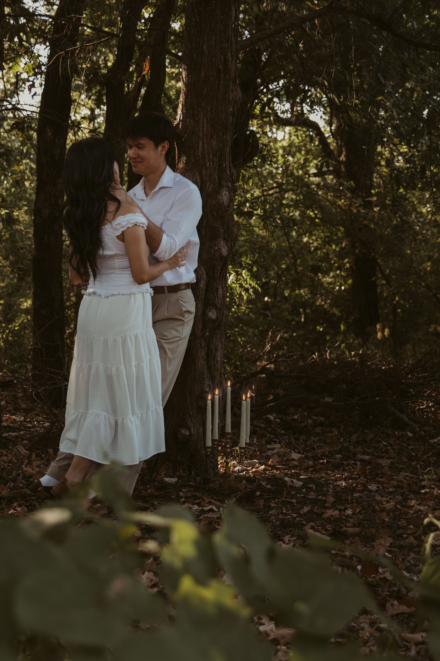 A romantic couple embraces in a wooded forest setting with dappled sunlight filtering through the trees.