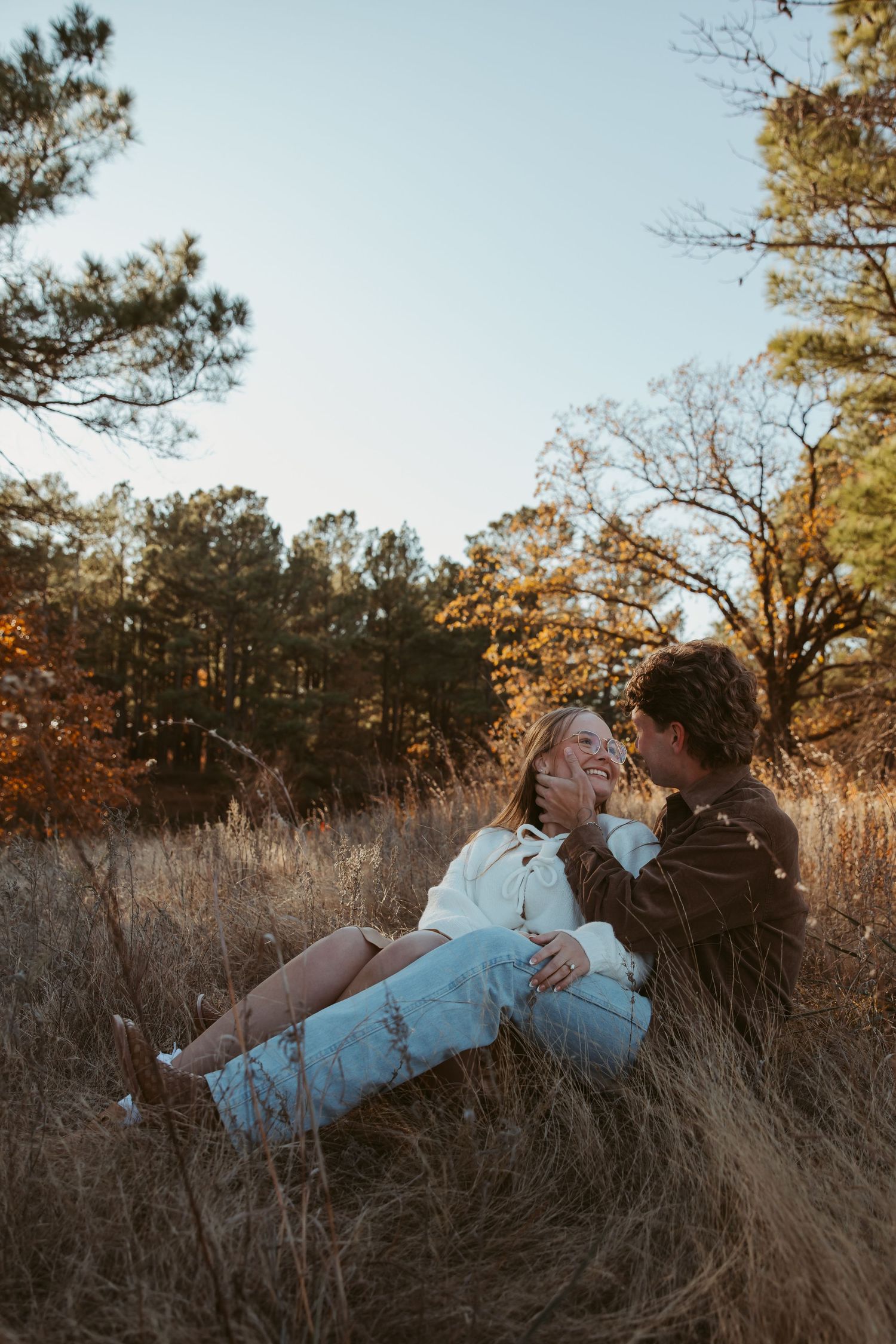 A romantic couple sits together in a golden field at sunset surrounded by autumn trees and tall grass. engagement photos DFW
