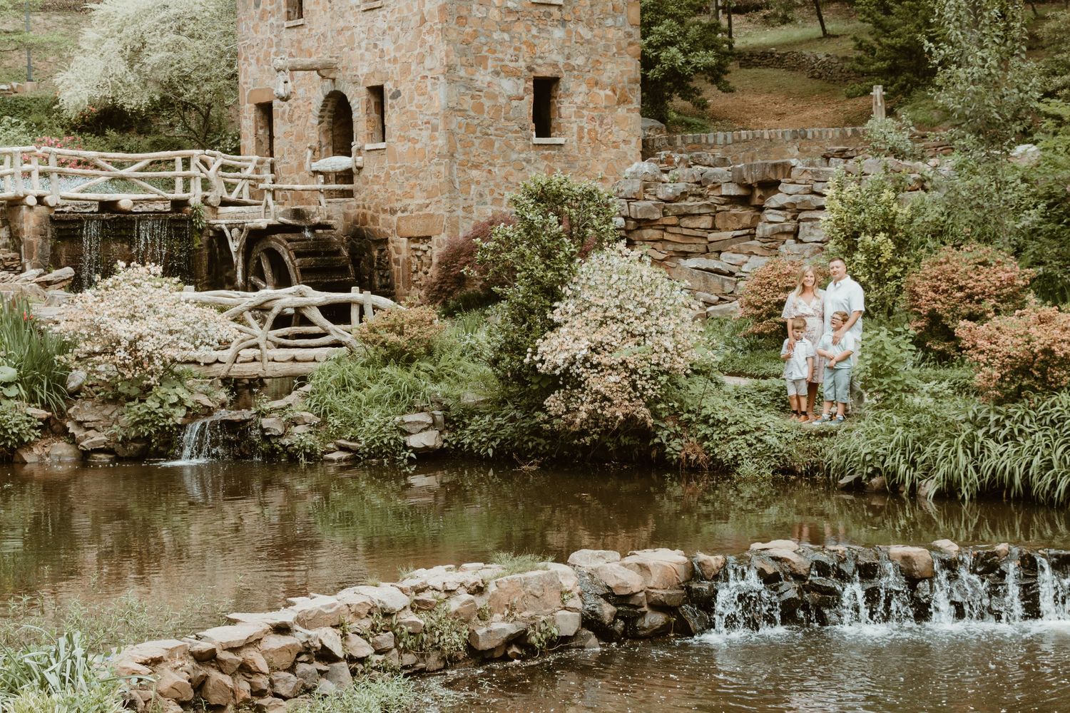 Historic stone mill building by a small waterfall with greenery surrounding the serene scene.