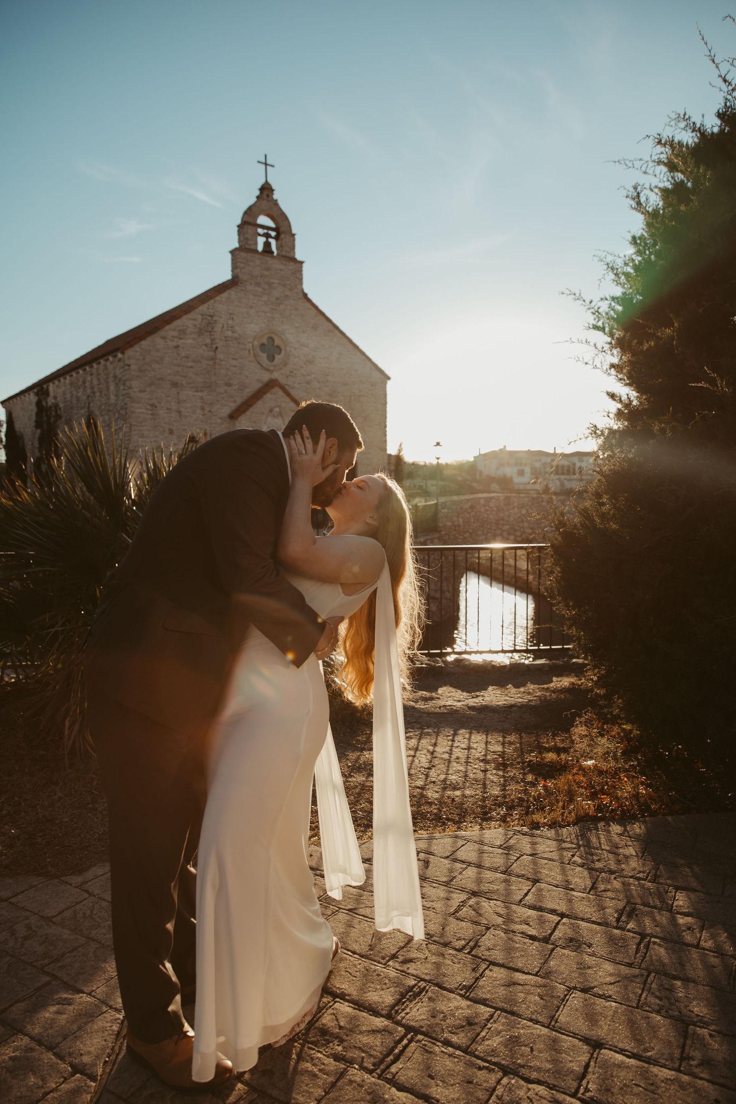 A romantic wedding couple embraces at sunset in front of a small stone chapel with a cross on top.