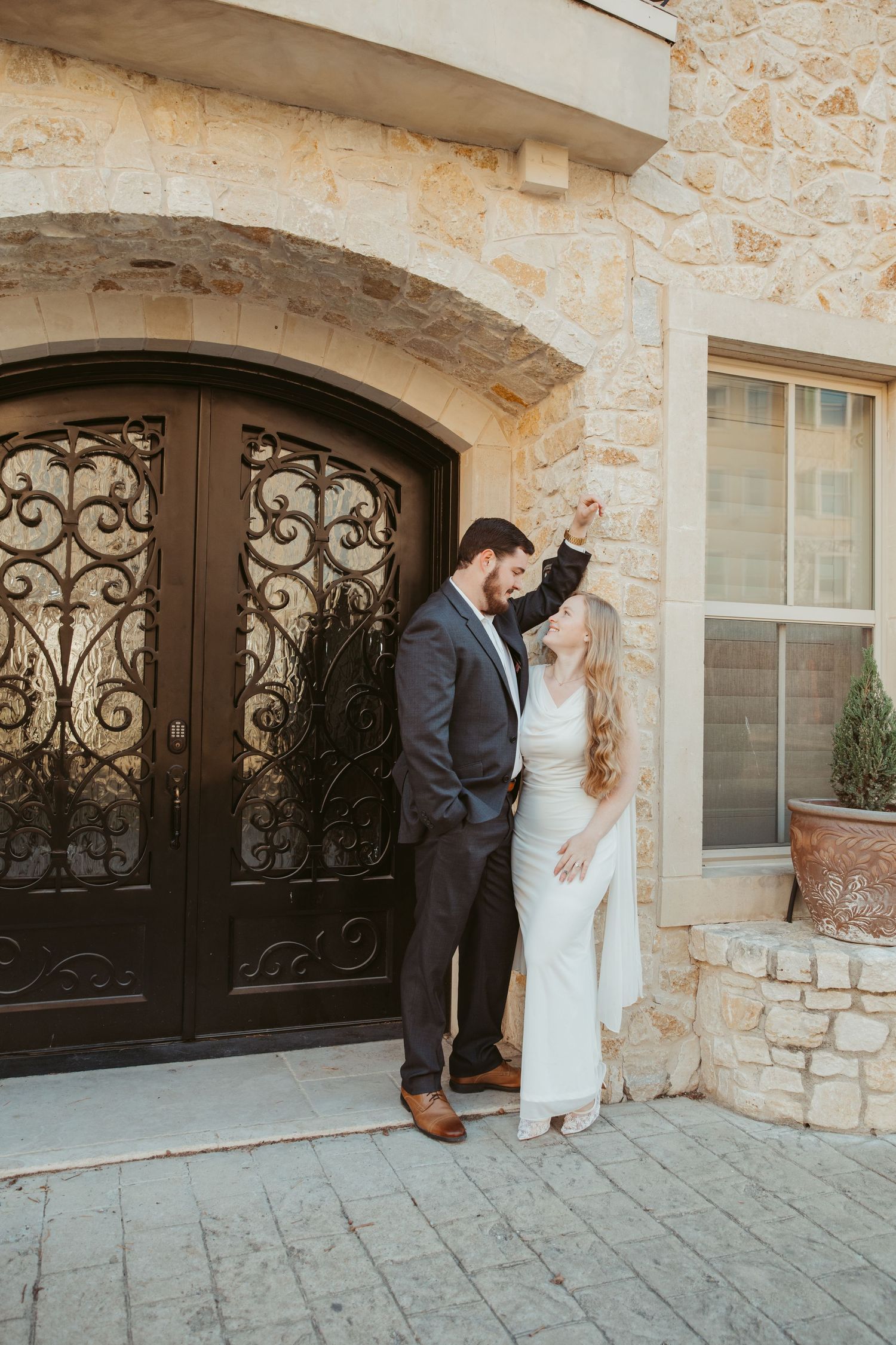 A couple in wedding attire share intimate moments against a stone wall with ornate iron window details.