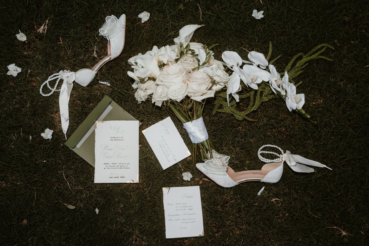 Elegant wedding flat lay with white peonies, invitation suite, and blush heels on green grass.