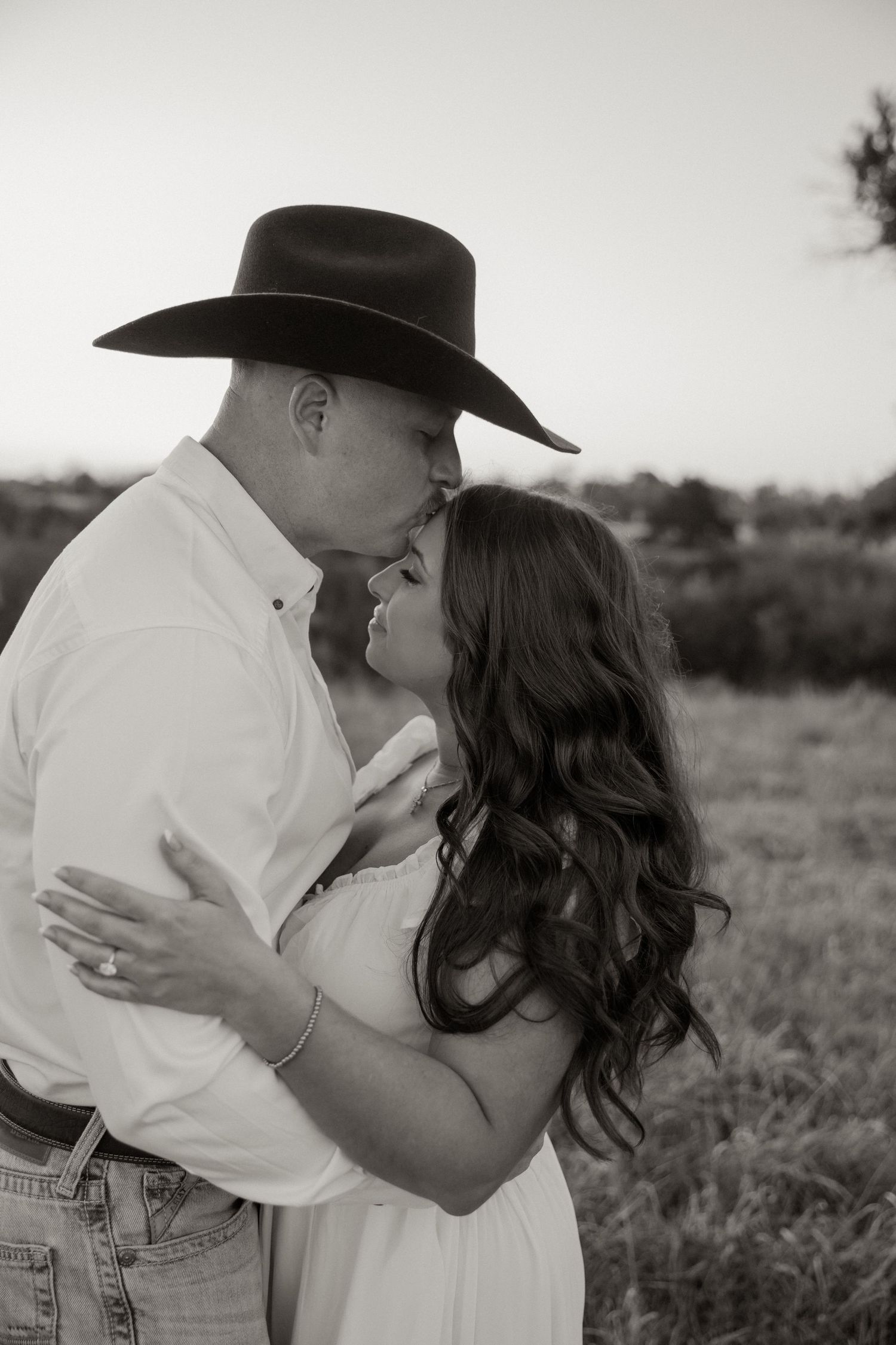 Black and white photo of a couple in western attire sharing a romantic moment in an outdoor field setting.