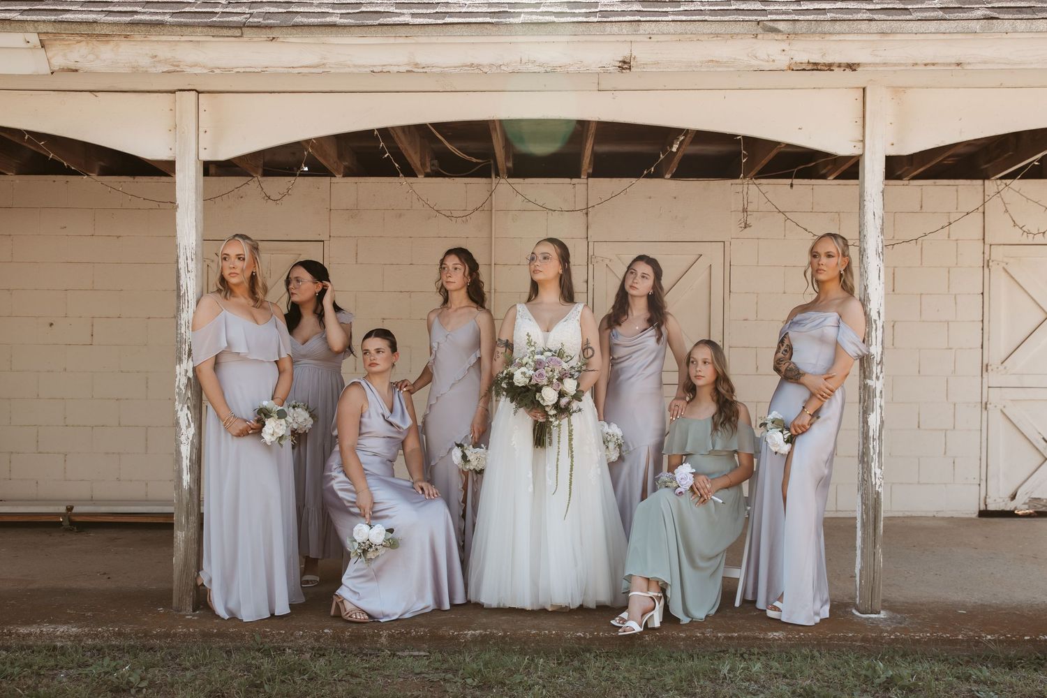 Bride in white gown poses with seven bridesmaids in lavender and sage dresses, holding bouquets outside a rustic barn.