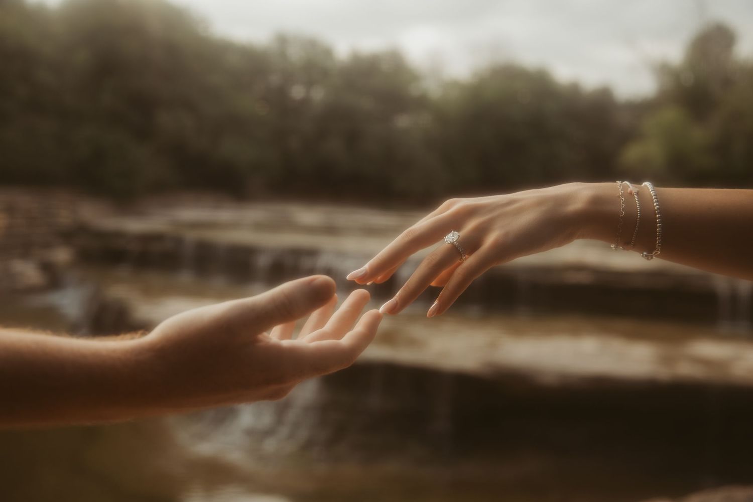 Two hands reaching toward each other near a serene river with rocky waterfall backdrop in soft natural light.