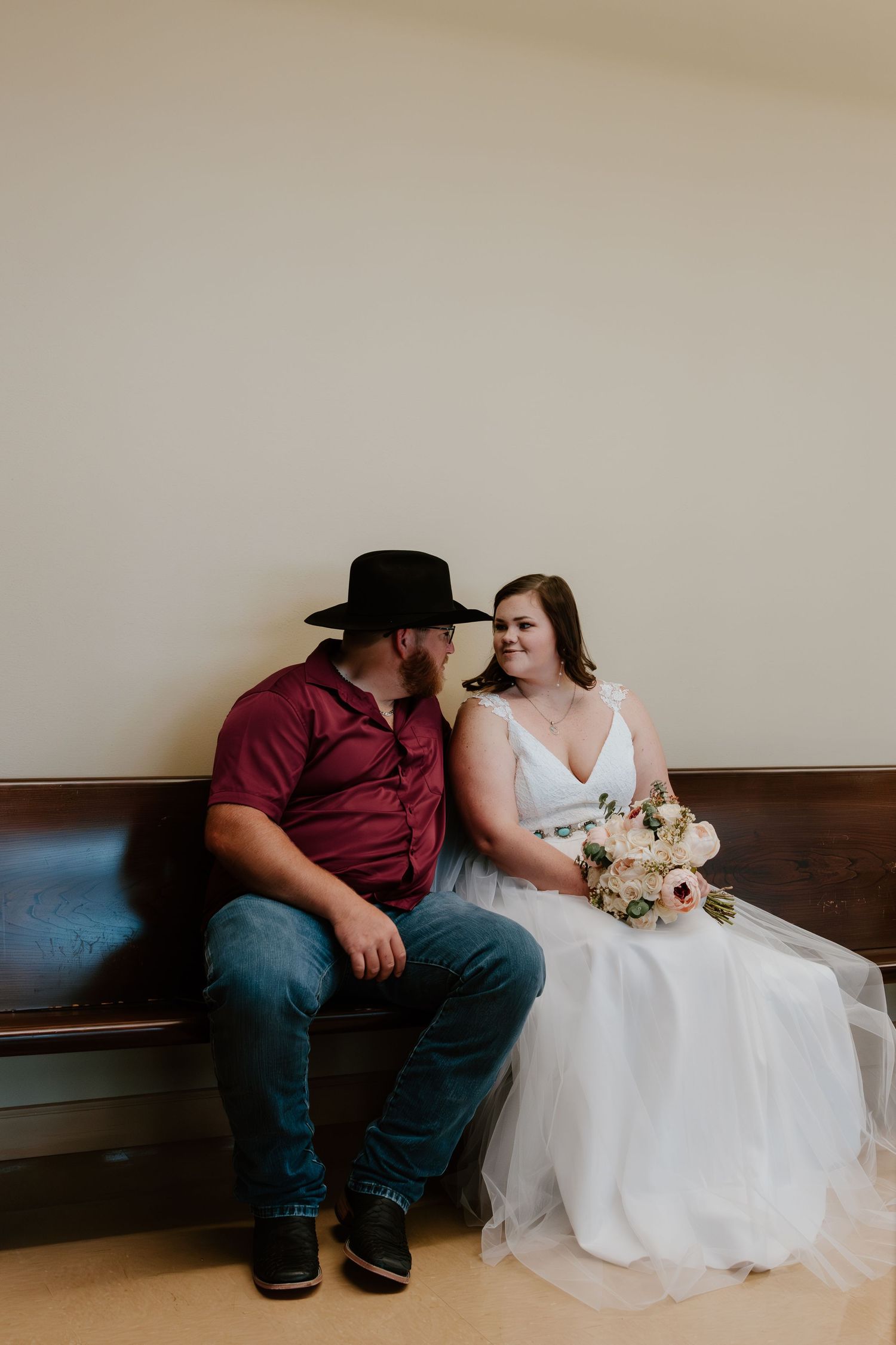A romantic wedding moment captured on a bench with a burgundy shirt, black hat, and flowing white dress.