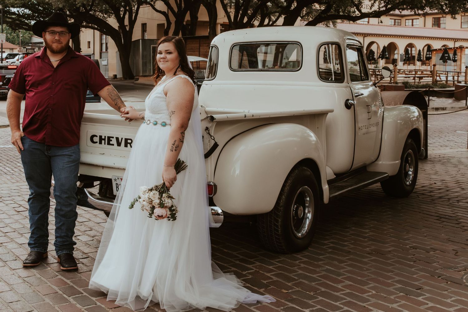 Newlyweds pose beside a vintage Chevrolet pickup truck on brick pavement.