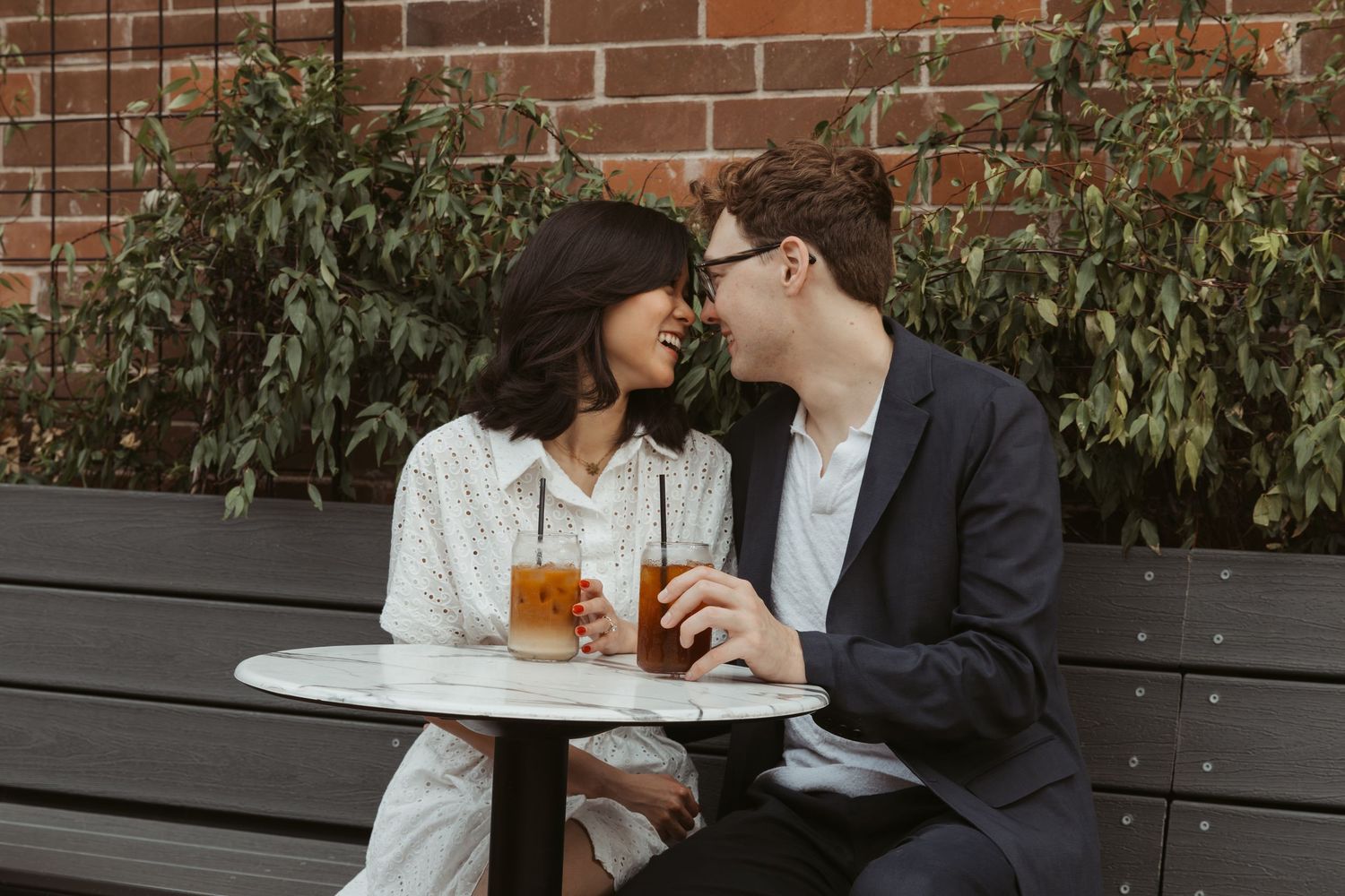 Two people sit at an outdoor cafe table with brick wall and greenery in the background, enjoying beverages together. Engagement photographer