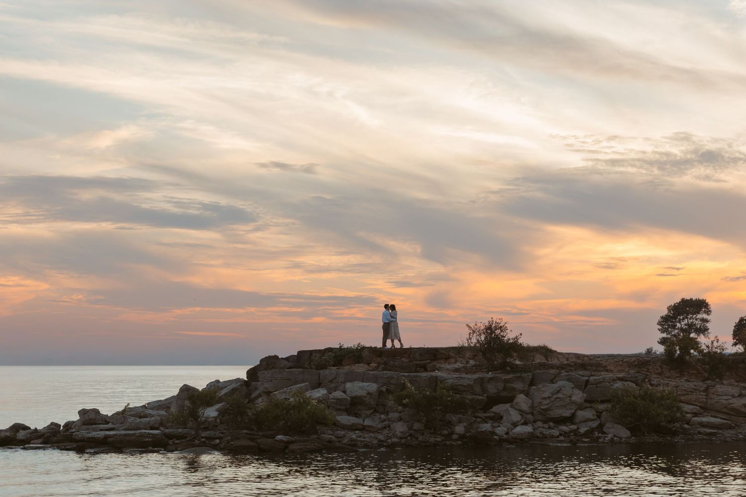 A panoramic view of a rocky coastline at sunset with dramatic clouds and silhouetted trees against an orange sky. Wedding photographer DFW