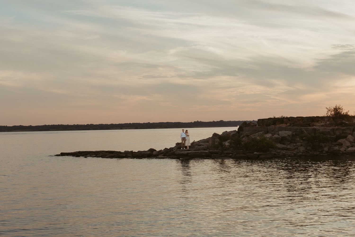 Rocky shoreline at sunset with calm waters and cloudy sky over a serene lake.