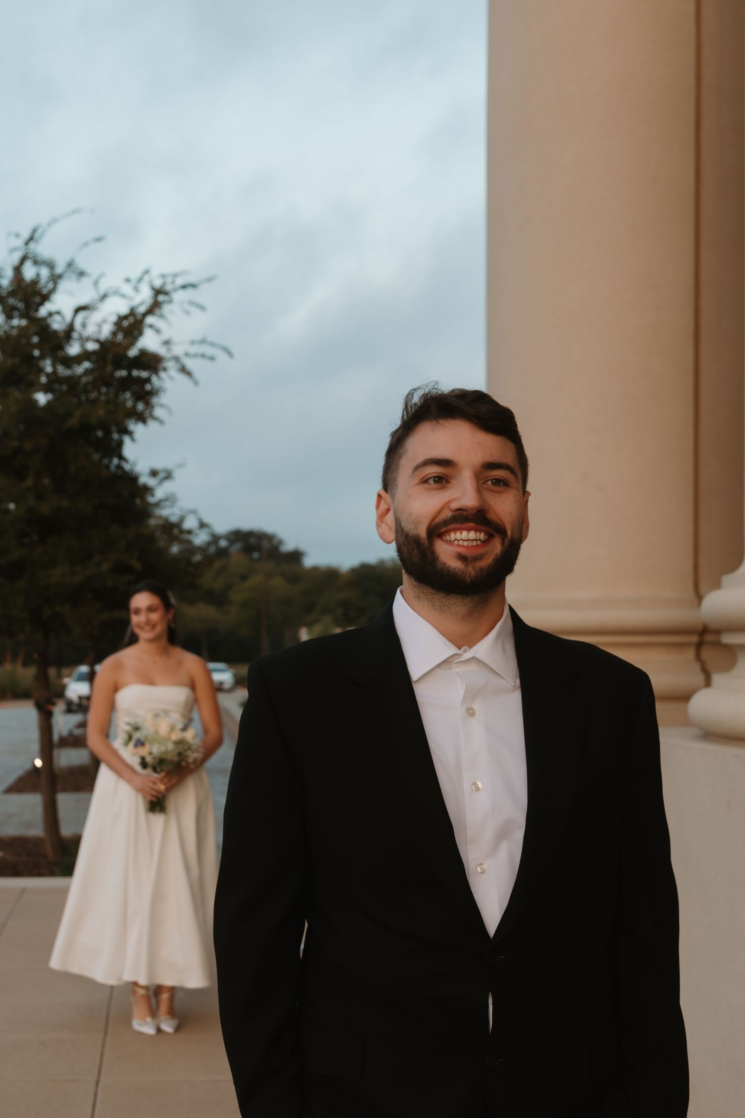 A wedding scene showing a person in a white dress and another in a black tuxedo standing by elegant white columns outdoors.