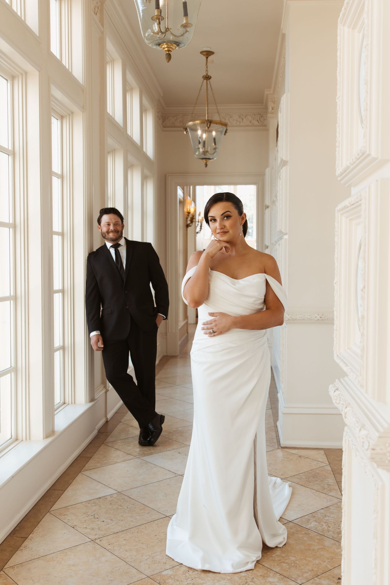 Elegant hallway with chandelier lighting and white walls features a romantic first look wedding moment.