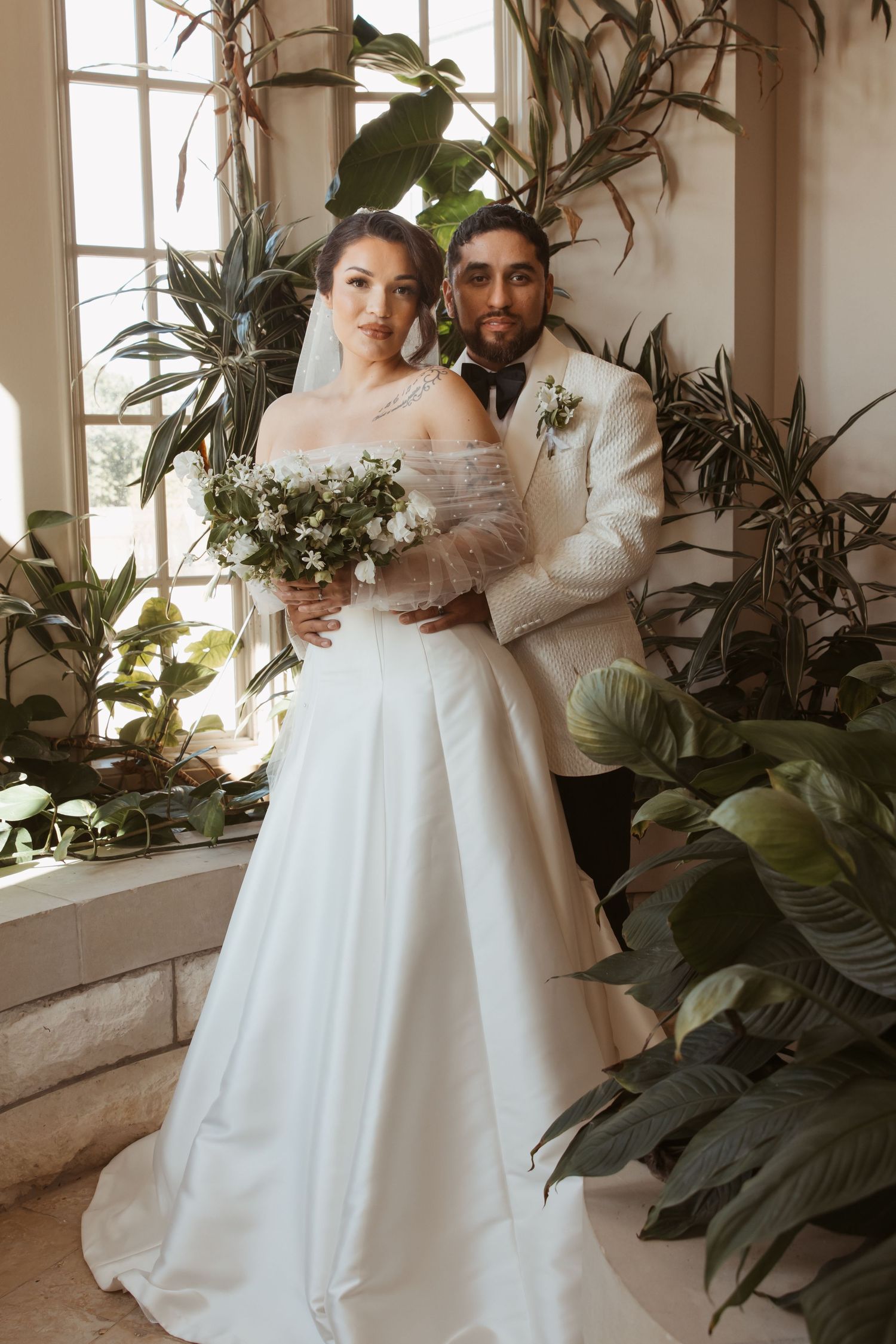 A couple poses together in a sunlit conservatory filled with tropical plants and palm fronds.