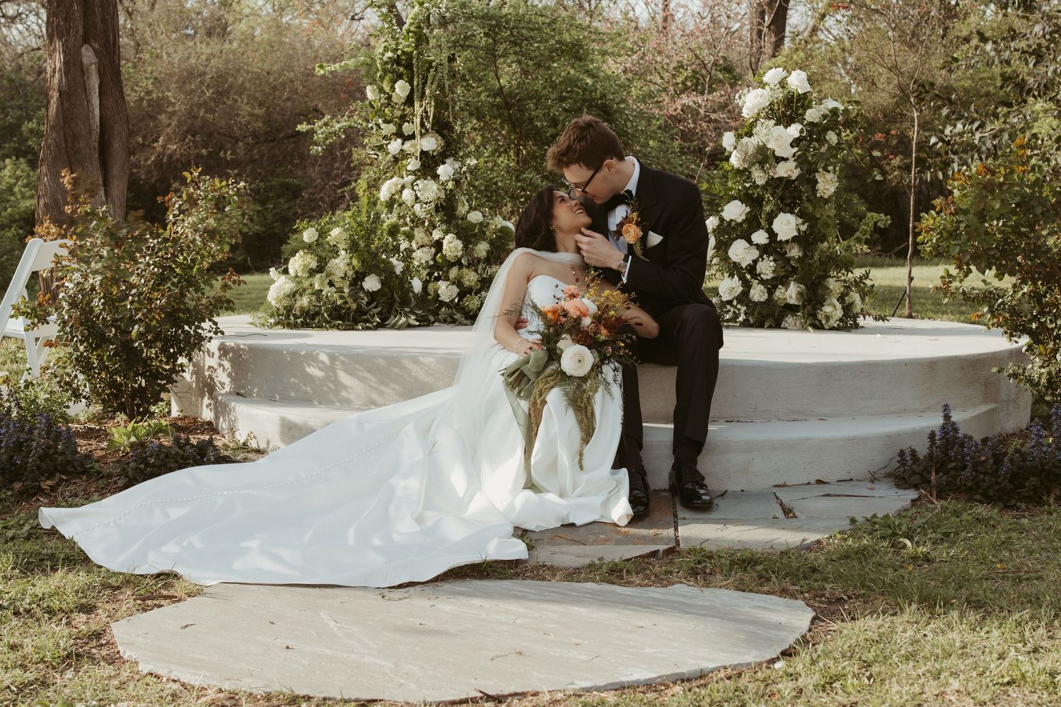 Bride and groom share romantic kiss beside white floral fountain in garden wedding setting.