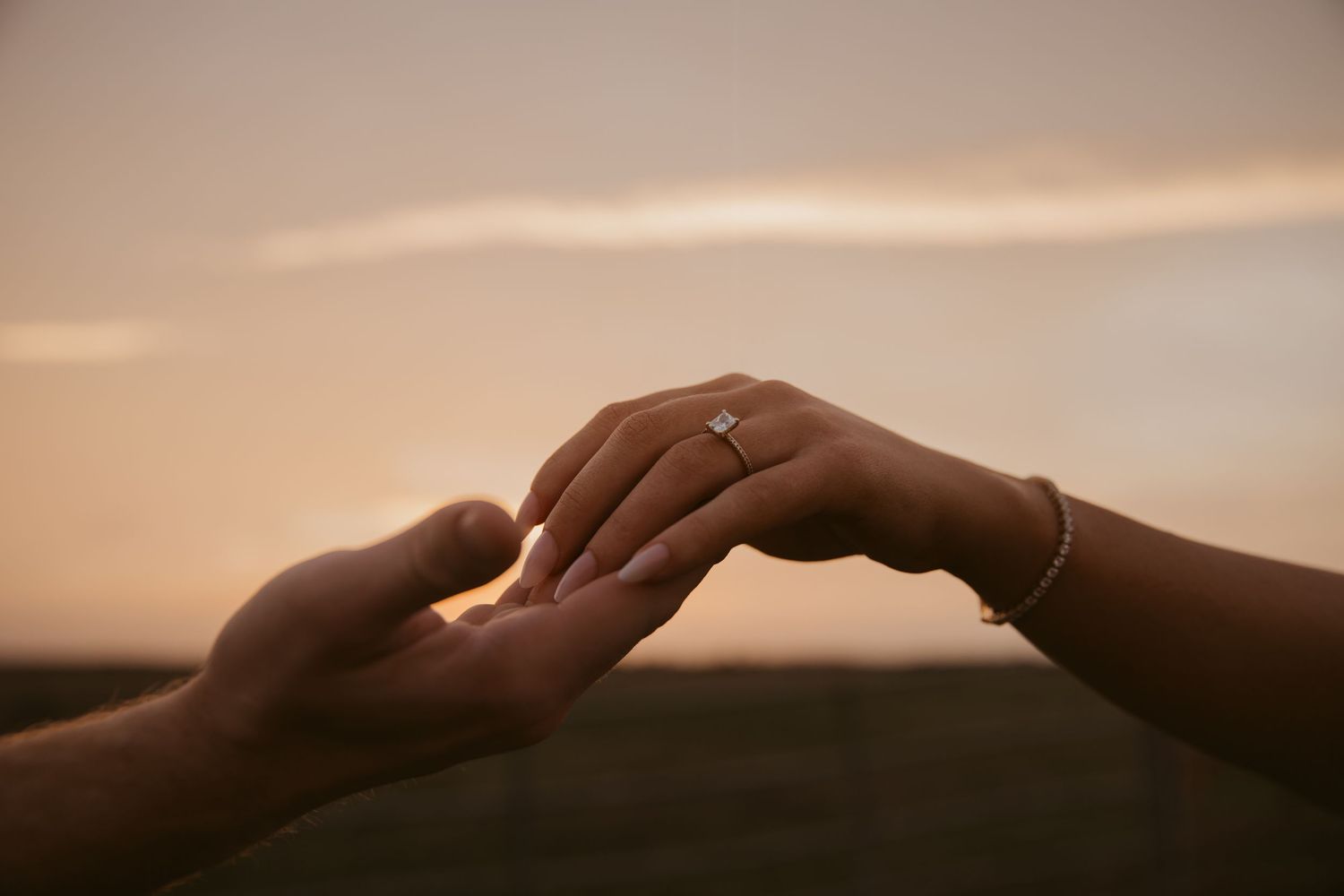 Two hands touching at sunset, one wearing a diamond engagement ring and bracelet, symbolizing a romantic proposal.