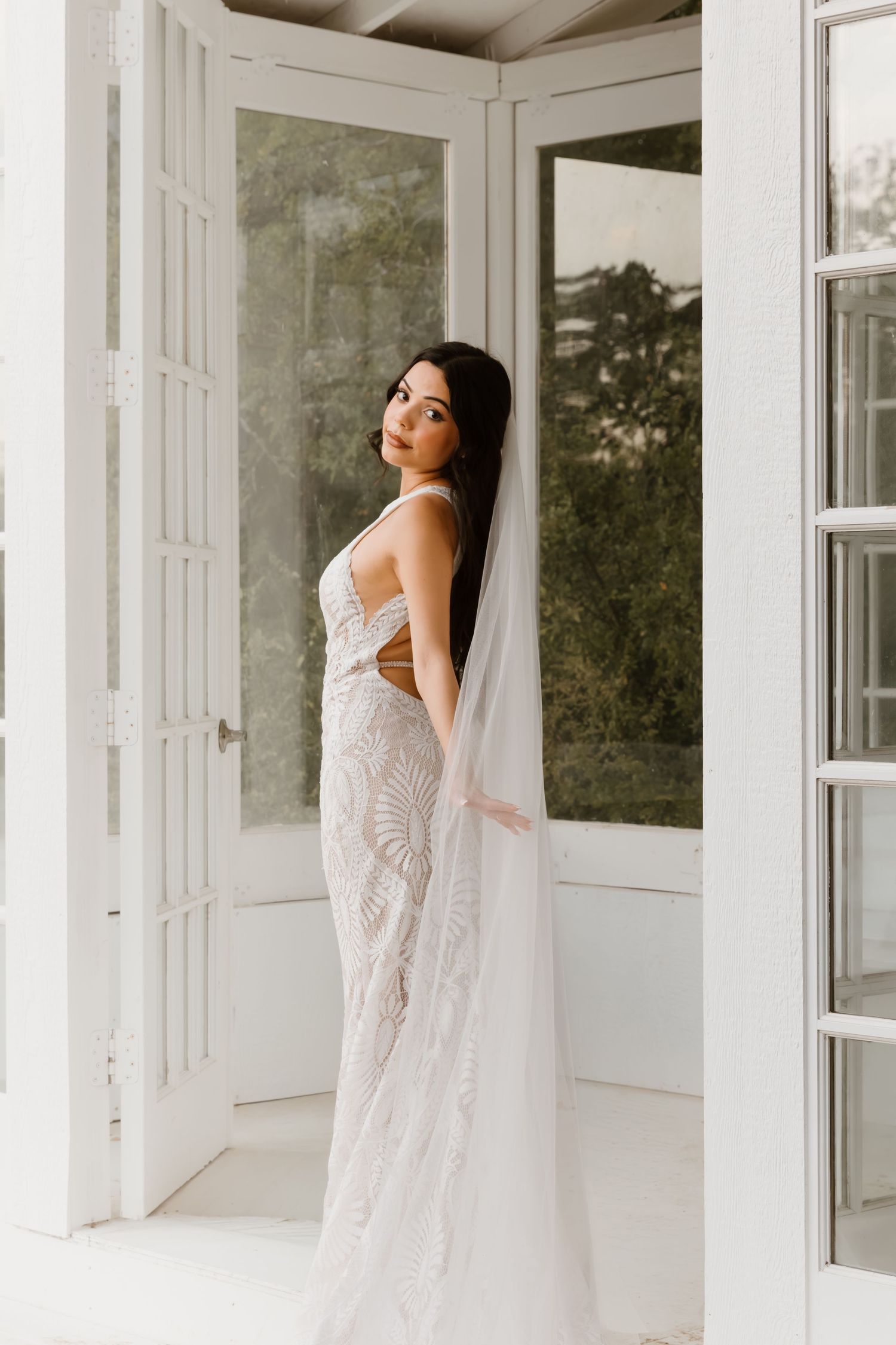 A bride in a flowing white lace wedding dress poses elegantly against white walls in a sunlit room.