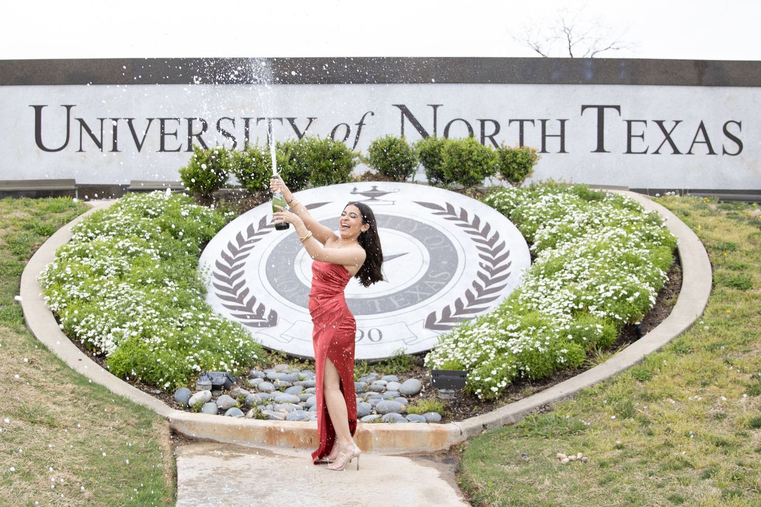 A person in red poses in front of a decorative University of North Texas sign surrounded by landscaped flowers.