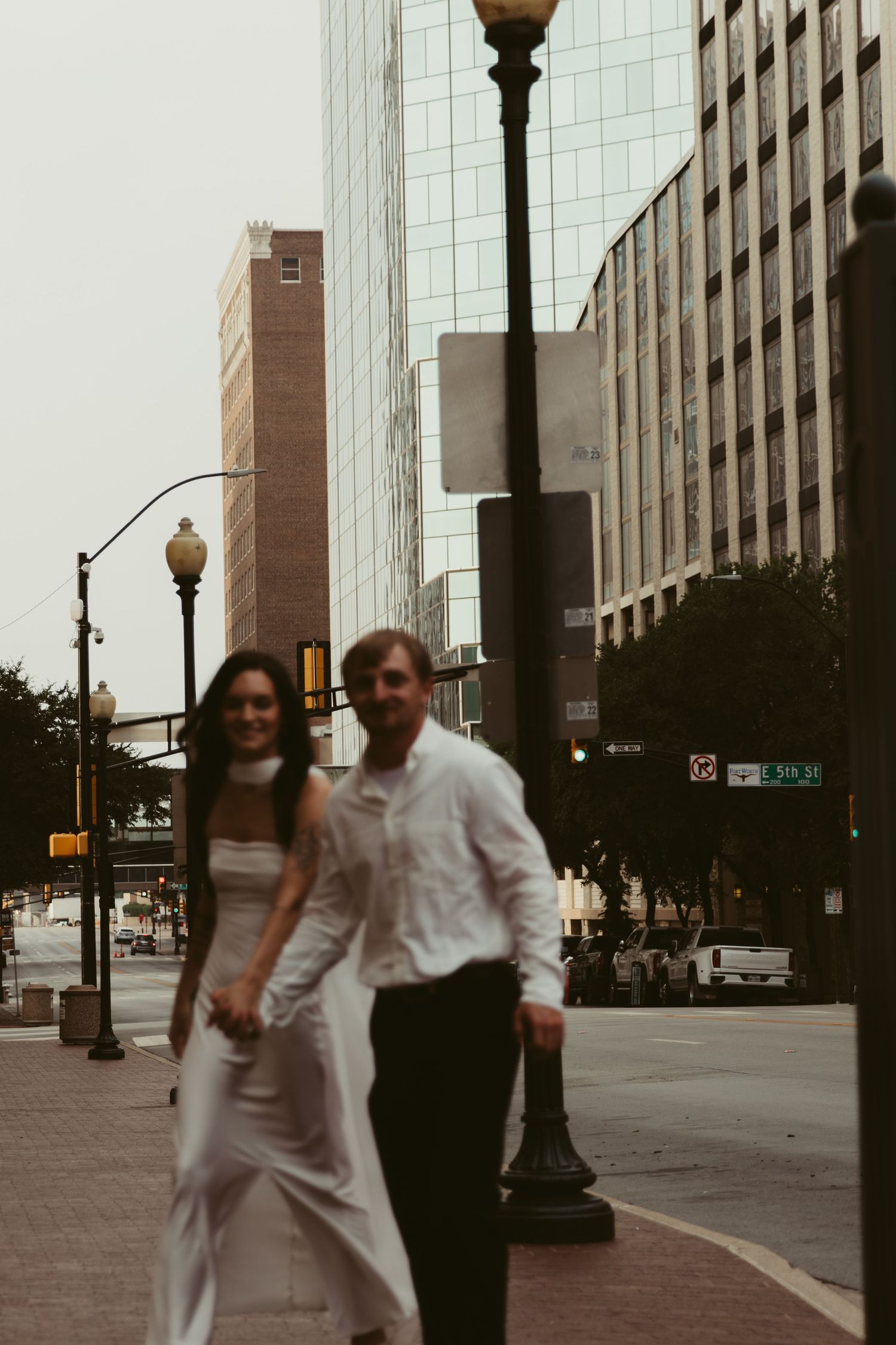 A couple in formal attire stroll together on a city sidewalk with downtown buildings and lamp posts in the background.