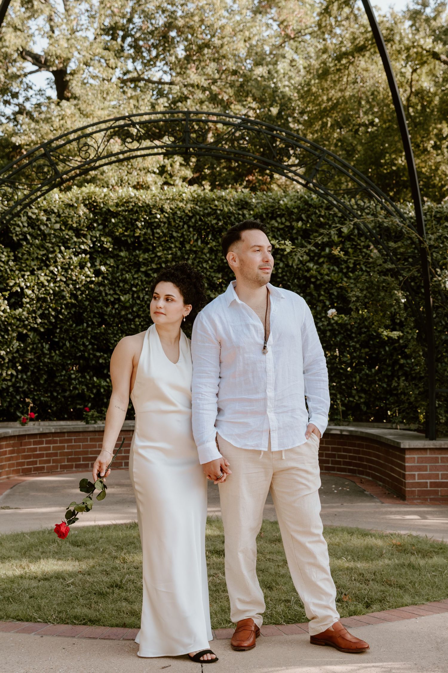A couple in matching white outfits holds hands while walking along a garden pathway in a romantic outdoor setting.