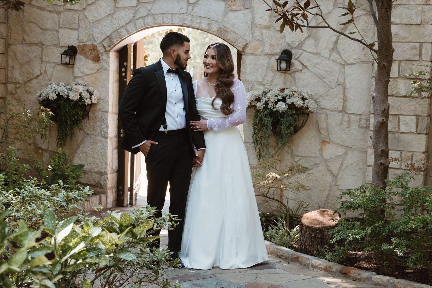 A romantic wedding couple stands in front of a stone archway decorated with elegant white floral arrangements.