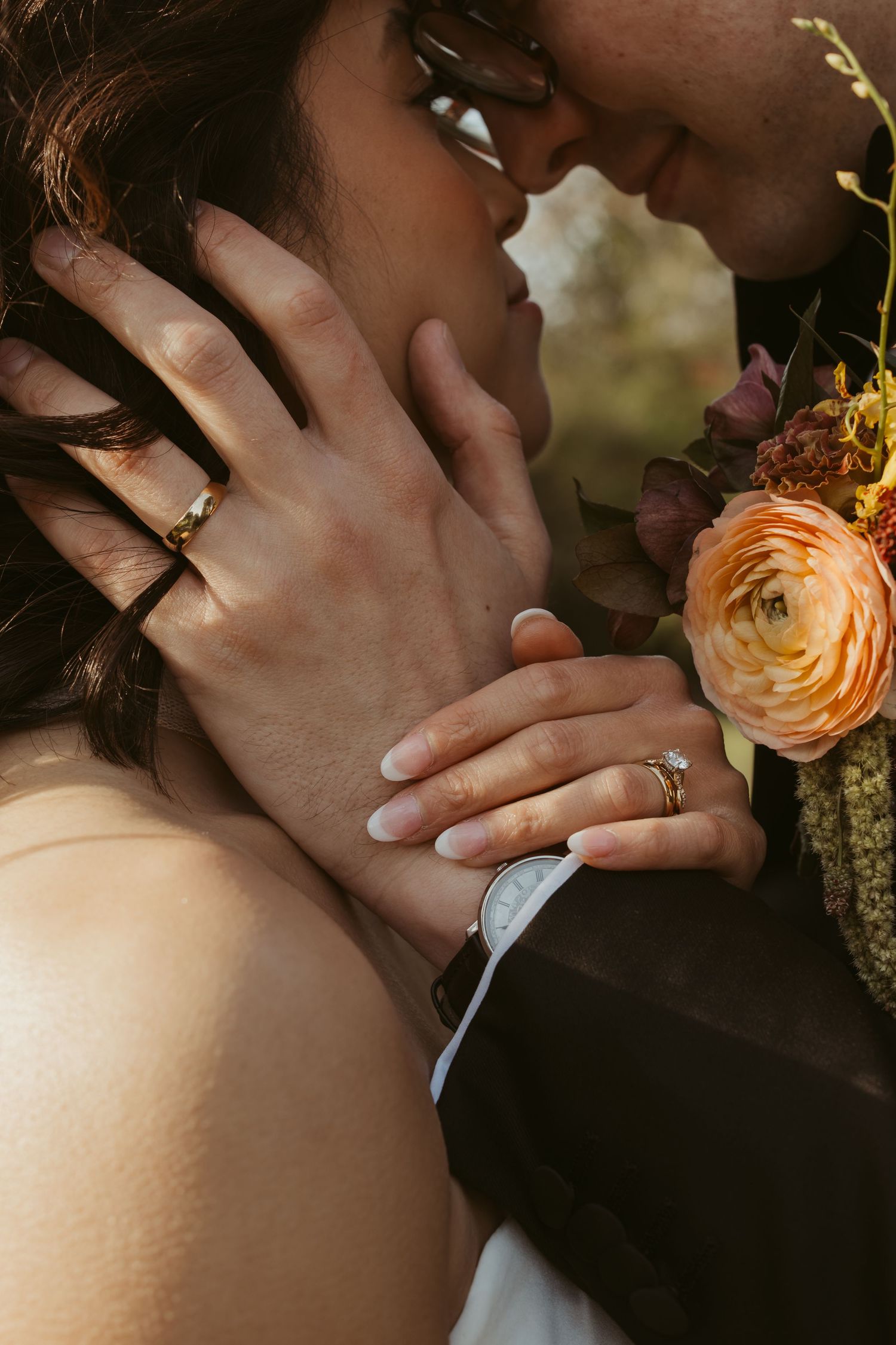 Close-up of newlyweds almost kissing, showcasing wedding rings and a boutonniere with orange ranunculus.