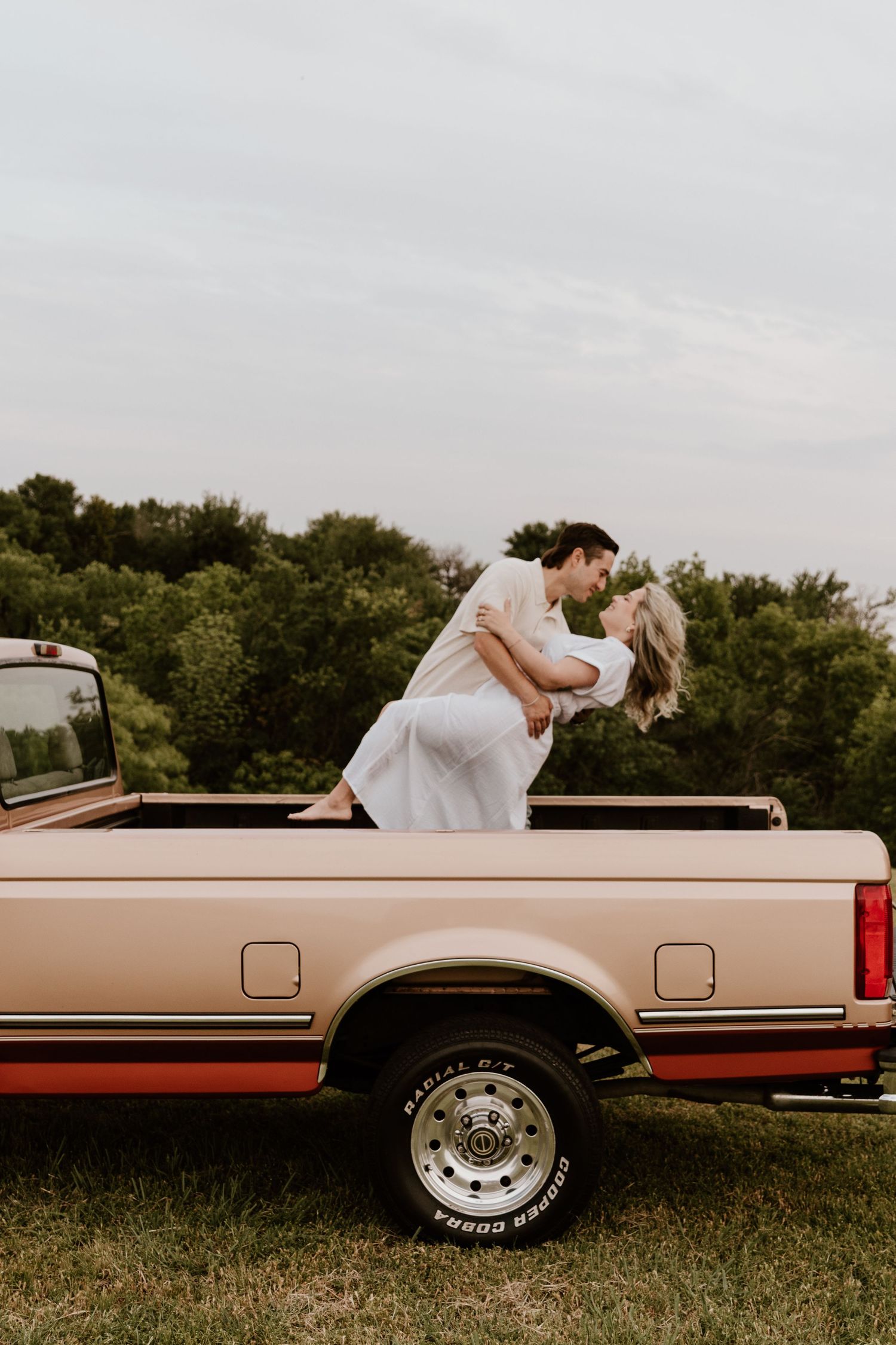 Romantic scene on vintage pickup truck with couple in white standing in truck bed against tree-lined backdrop at sunset.