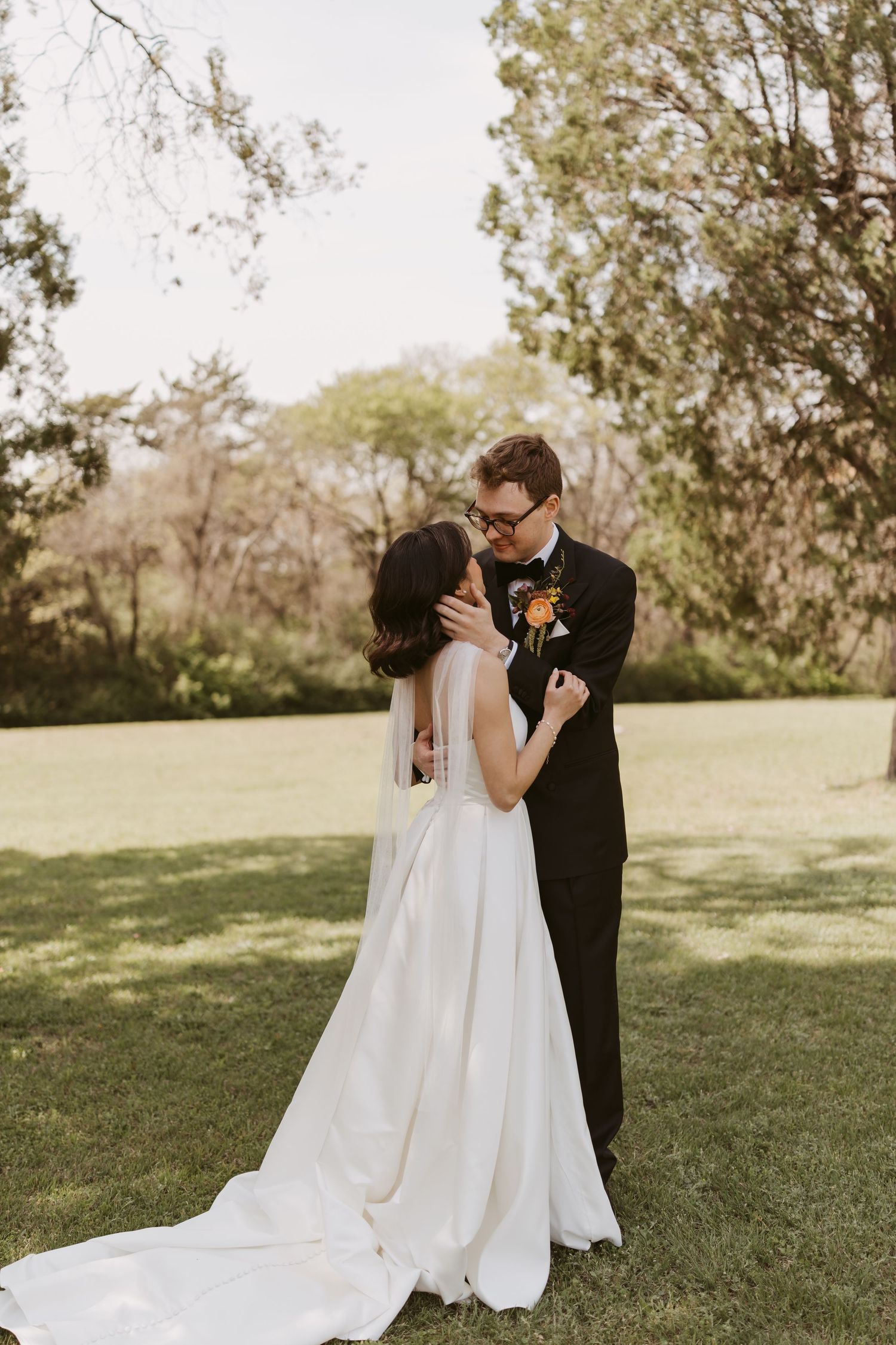 Bride and groom share first kiss on green lawn surrounded by trees in outdoor wedding ceremony.