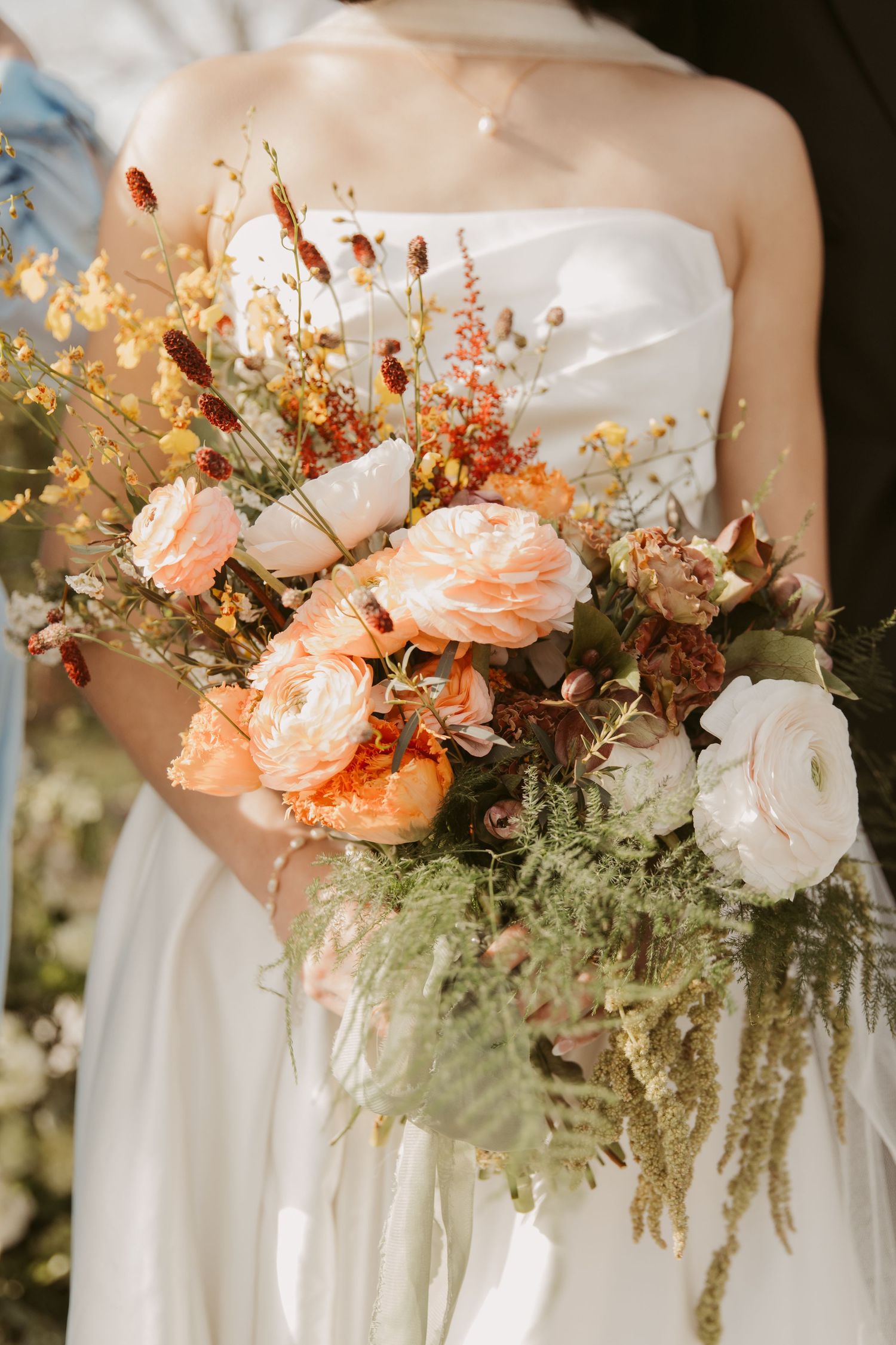 Bride in white strapless gown holding lush bridal bouquet with peach, white, and rust flowers with wild greenery.