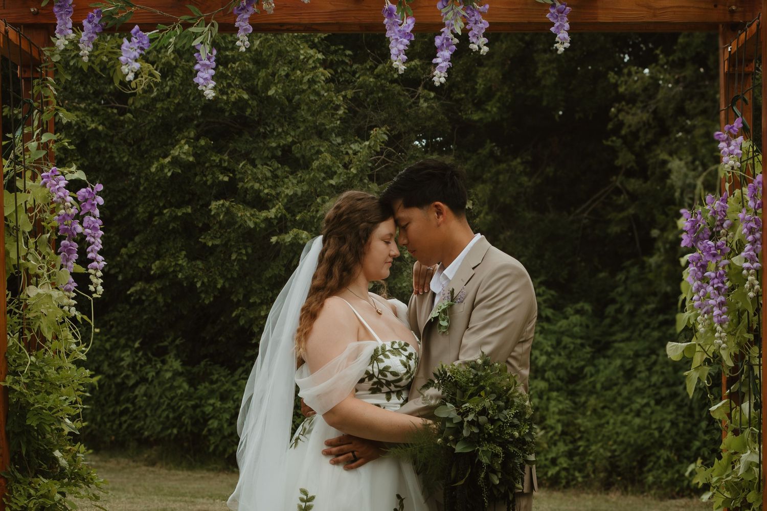 A romantic outdoor wedding ceremony under a wooden arch decorated with hanging purple wisteria flowers and greenery.