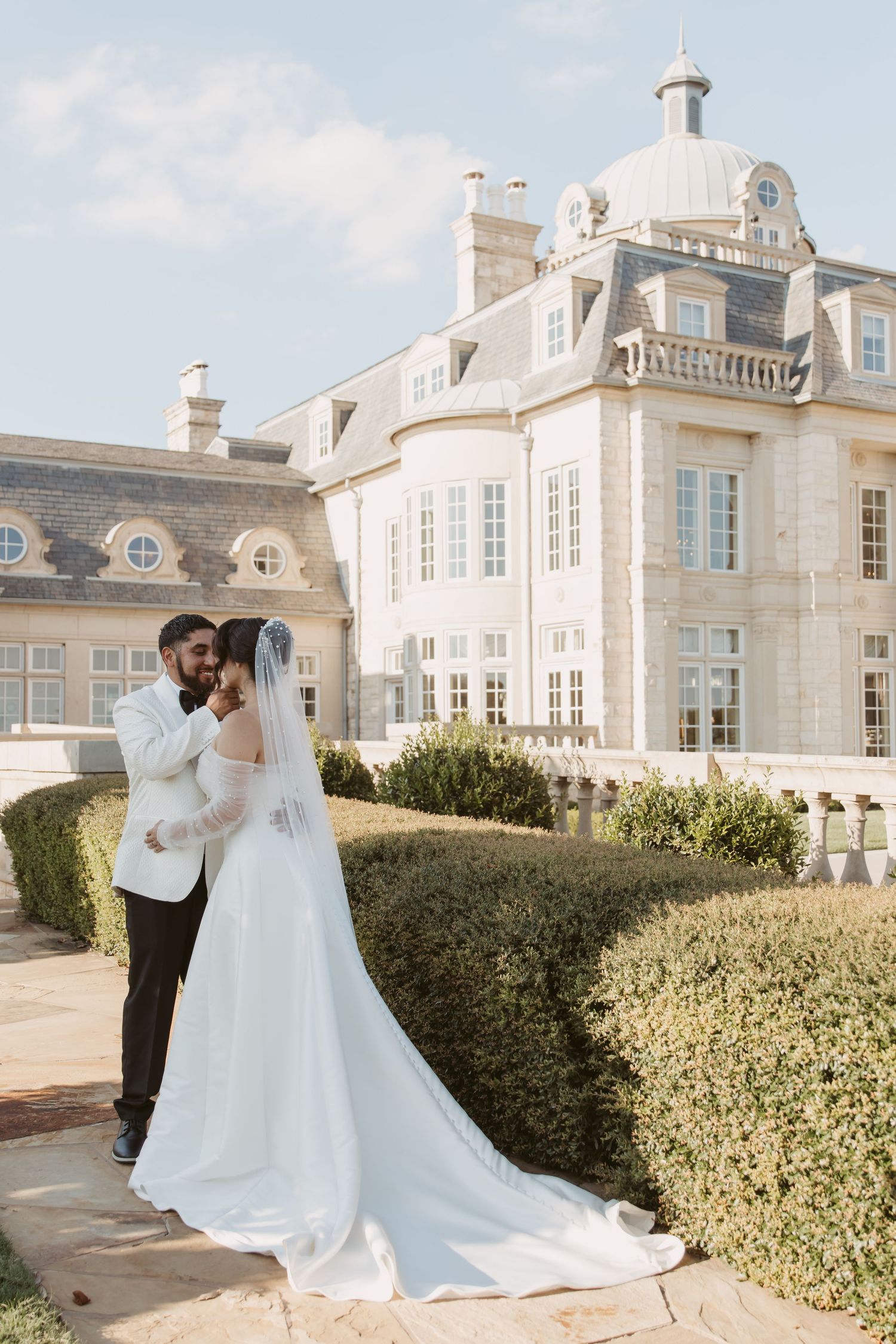 A romantic wedding couple shares intimate moments in front of an elegant white mansion with manicured hedges.