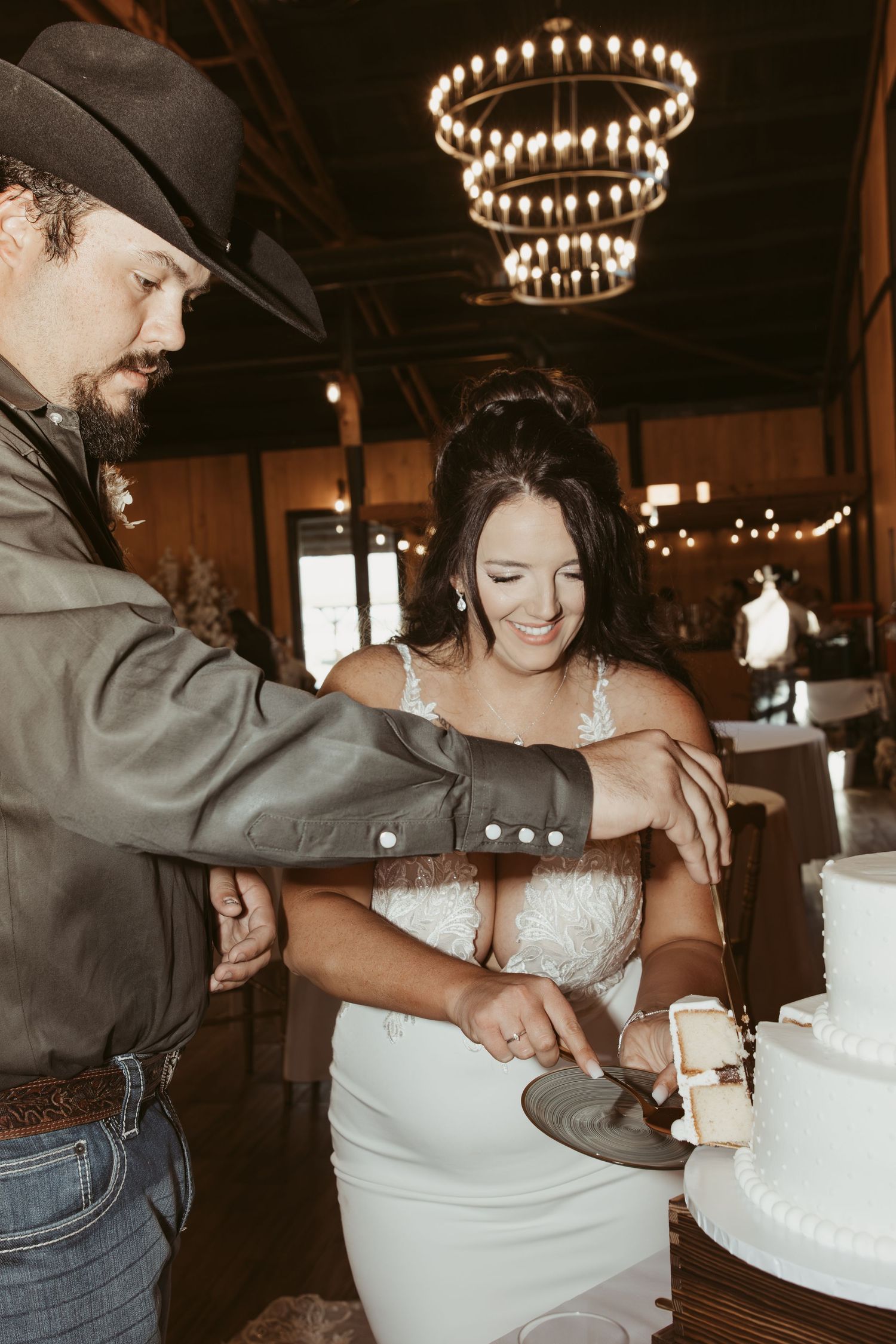 Wedding couple cutting their white tiered cake under rustic chandelier lighting. Wedding photographer DFW