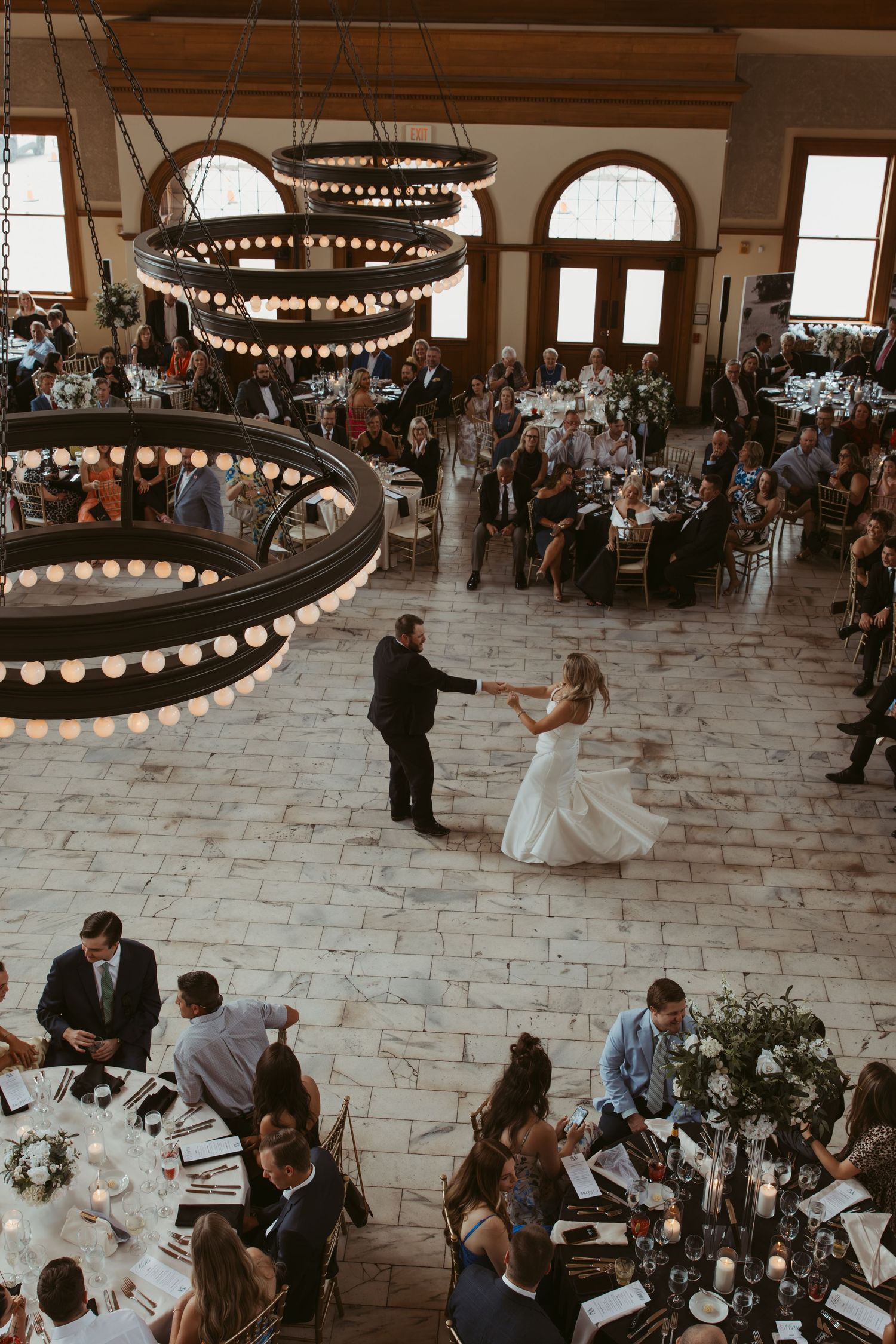 A couple dances beneath elegant circular chandeliers during a wedding reception in a grand event space with stone floors.