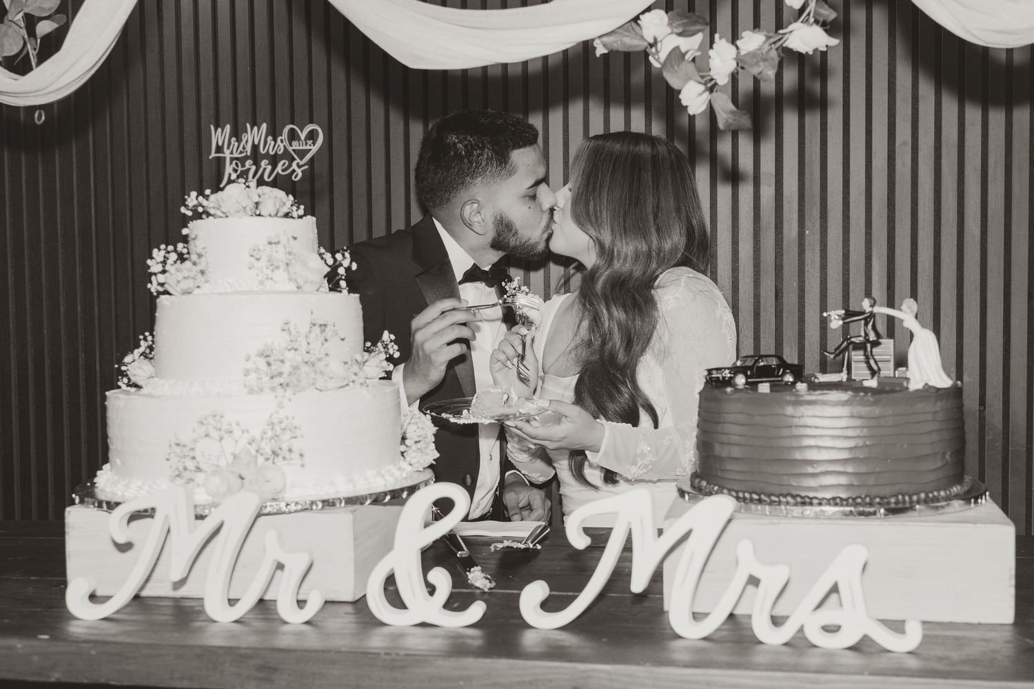 A black and white photo of newlyweds sharing a moment at their wedding reception with Mr & Mrs sign and tiered cakes. Wedding Photographer