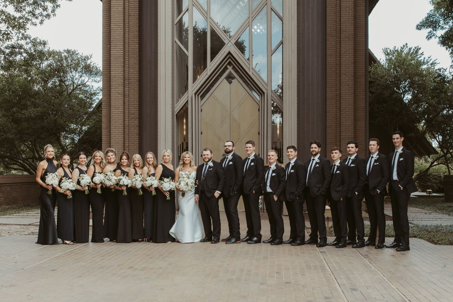 Wedding party poses together in formal attire outside a modern church building with glass windows and stone columns. Wedding Photographer DF