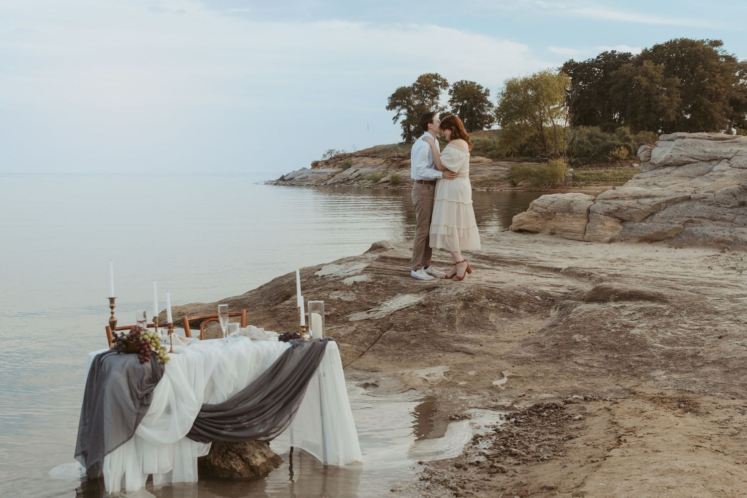 Elegant waterfront setup with draped table and couple on rocky shore.