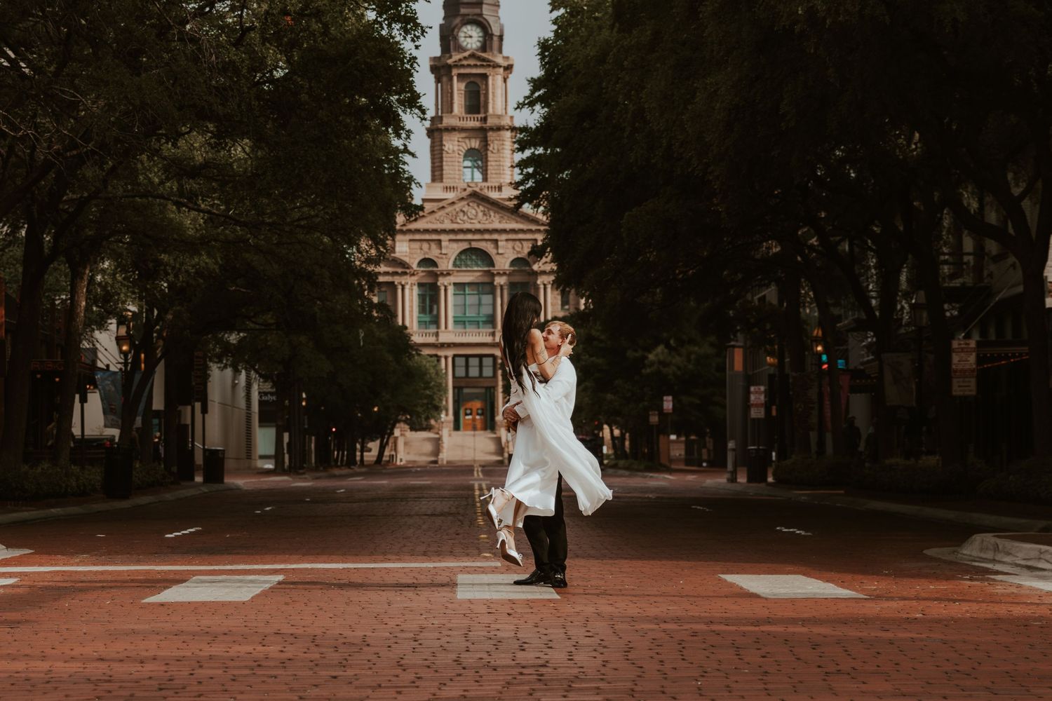 A couple in formal wedding attire poses in front of a historic brick building and tree-lined path. Wedding Photographer DFW