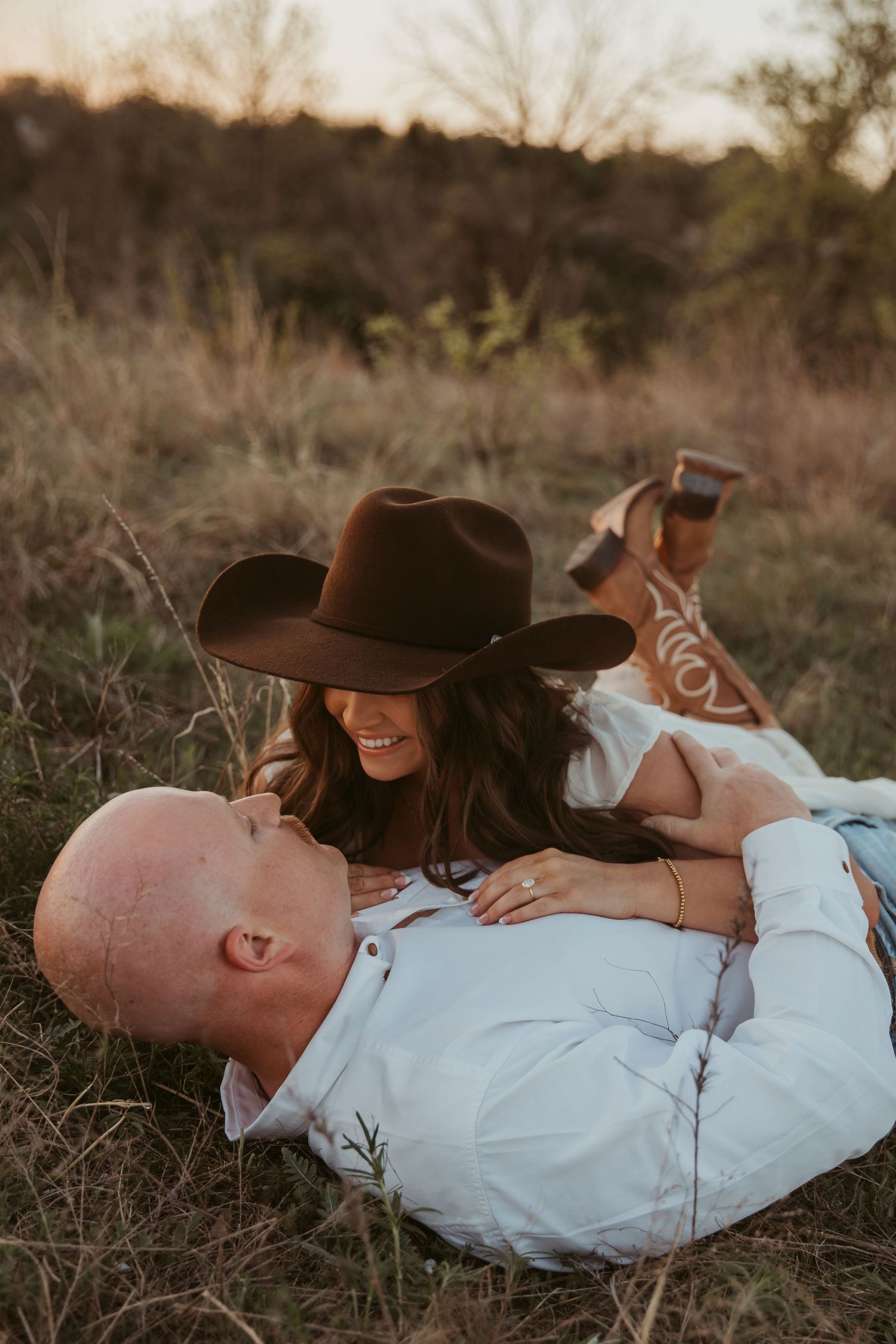 A couple relaxes in a golden field, woman in cowboy hat and boots smiling at bald man lying in her lap.