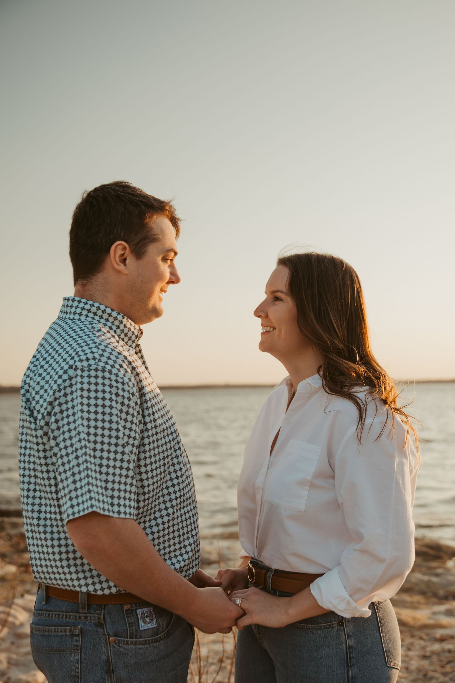 Couple holding hands and smiling at each other on a beach at golden hour sunset.