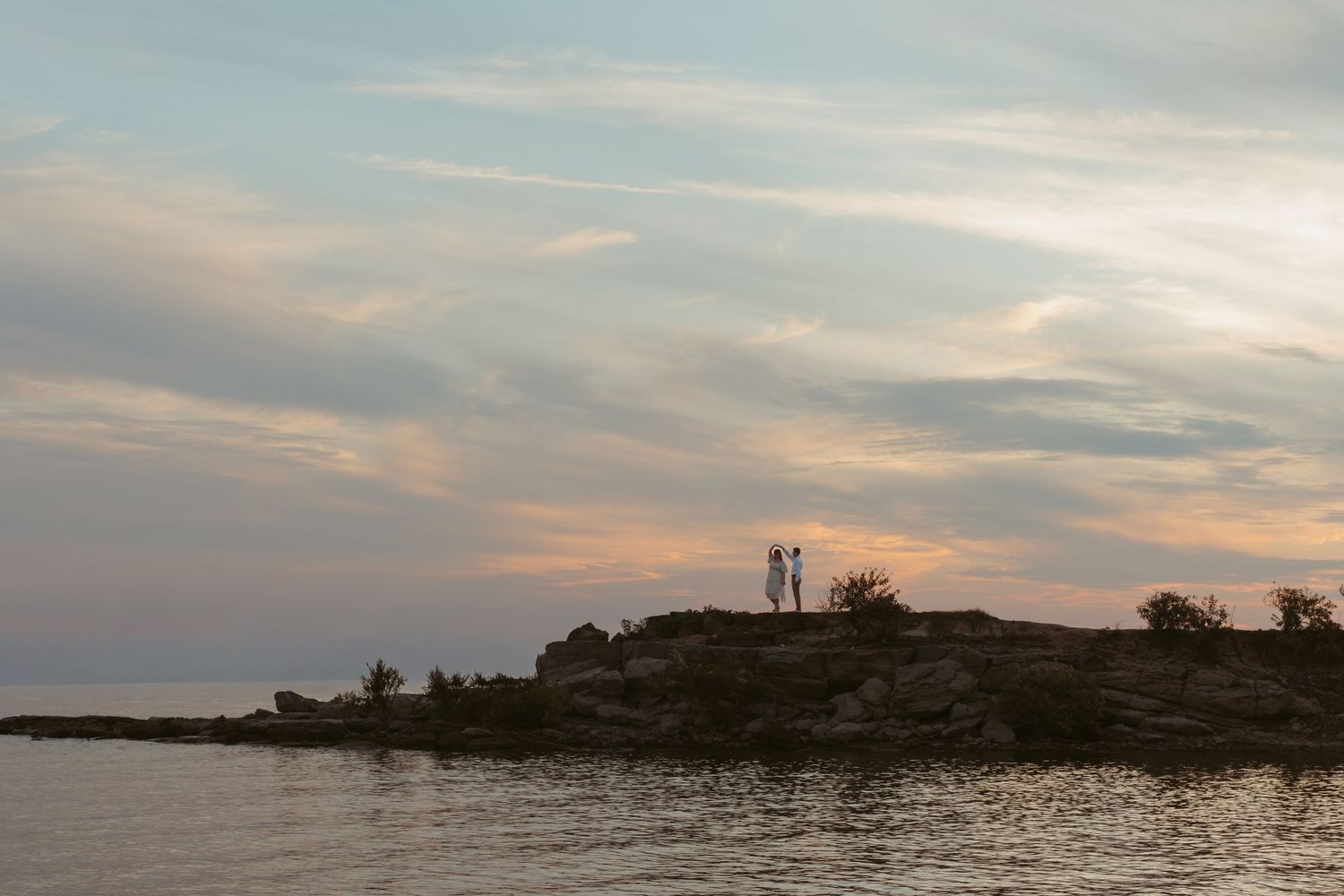 Silhouetted figures stand on a rocky coastal outcrop at sunset with dramatic cloudy skies and ocean views.